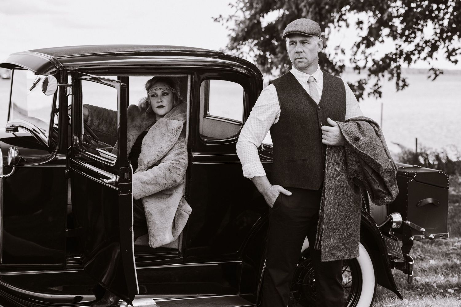A vintage black and white photograph showing people with a classic car from the early 20th century.