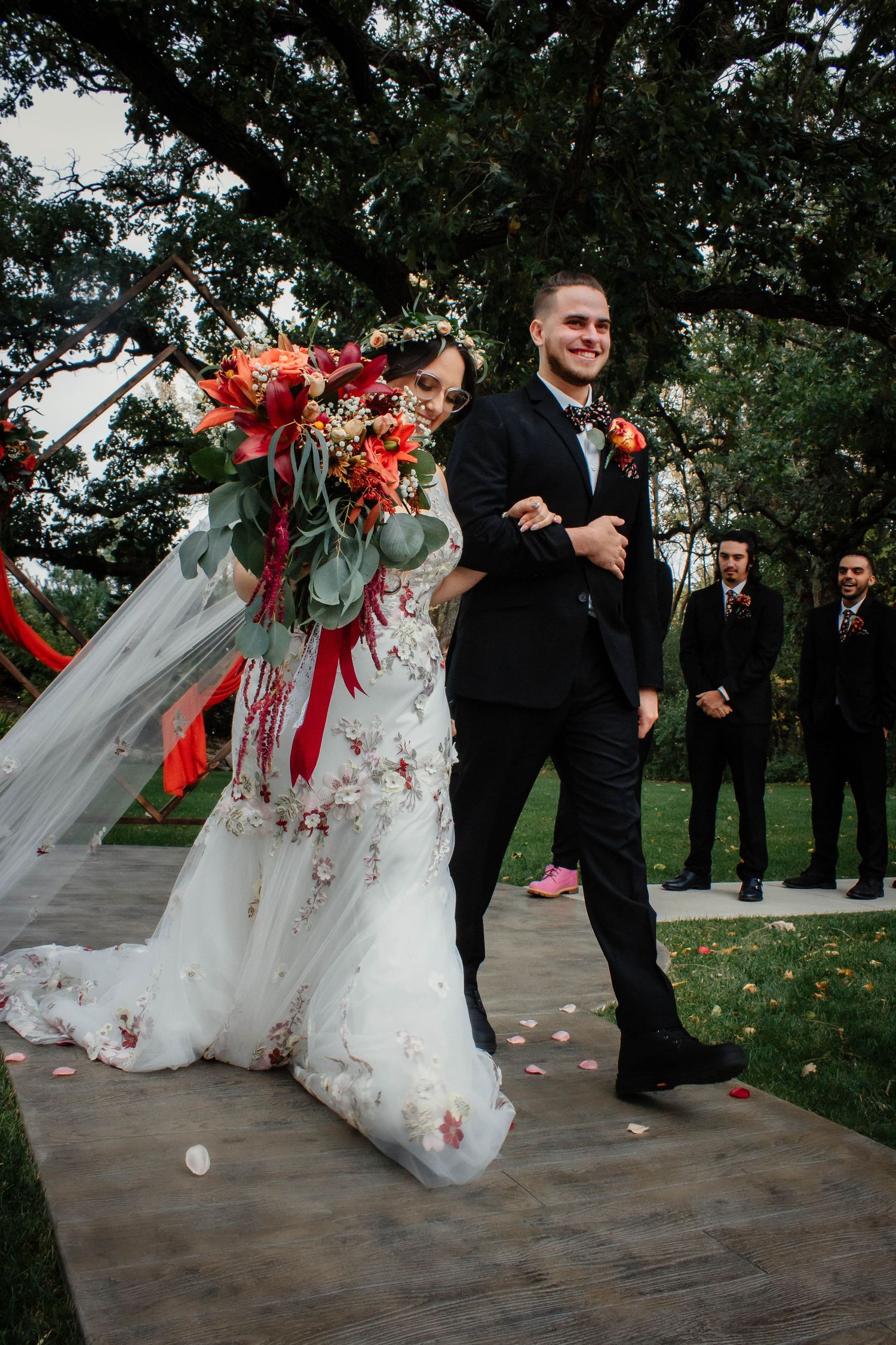 Wedding party dancing joyfully on an outdoor patio with red floral bouquets.