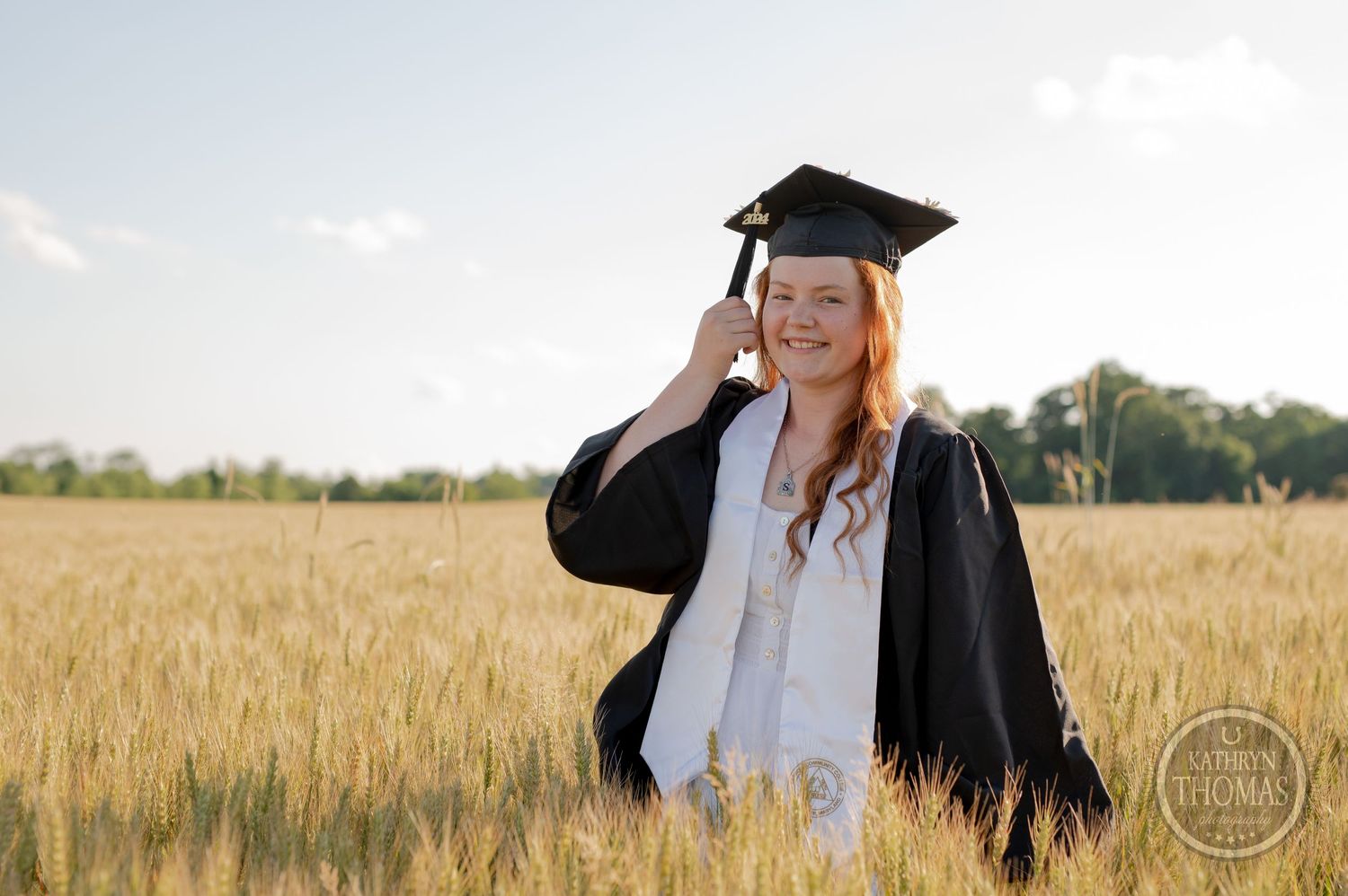 Graduate adjusts mortarboard while standing in wheat field during sunset.