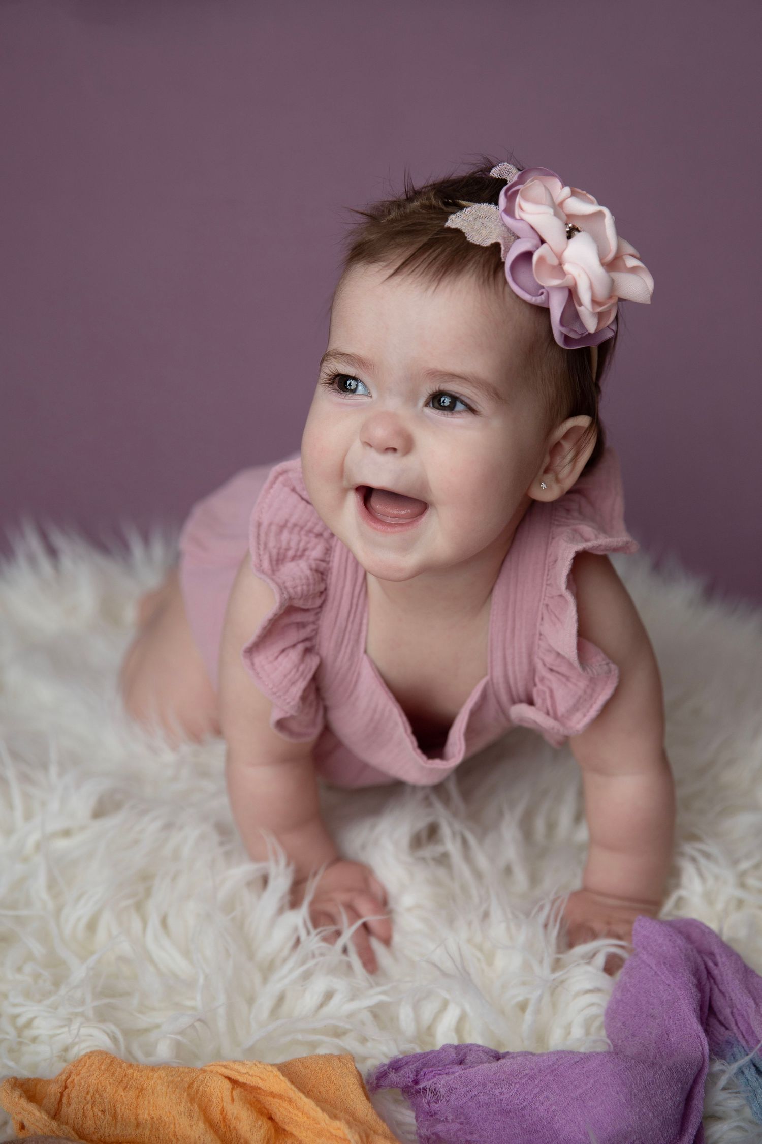 Happy smiling baby girl in pink ruffle romper with floral headband crawling on white fluffy rug.
