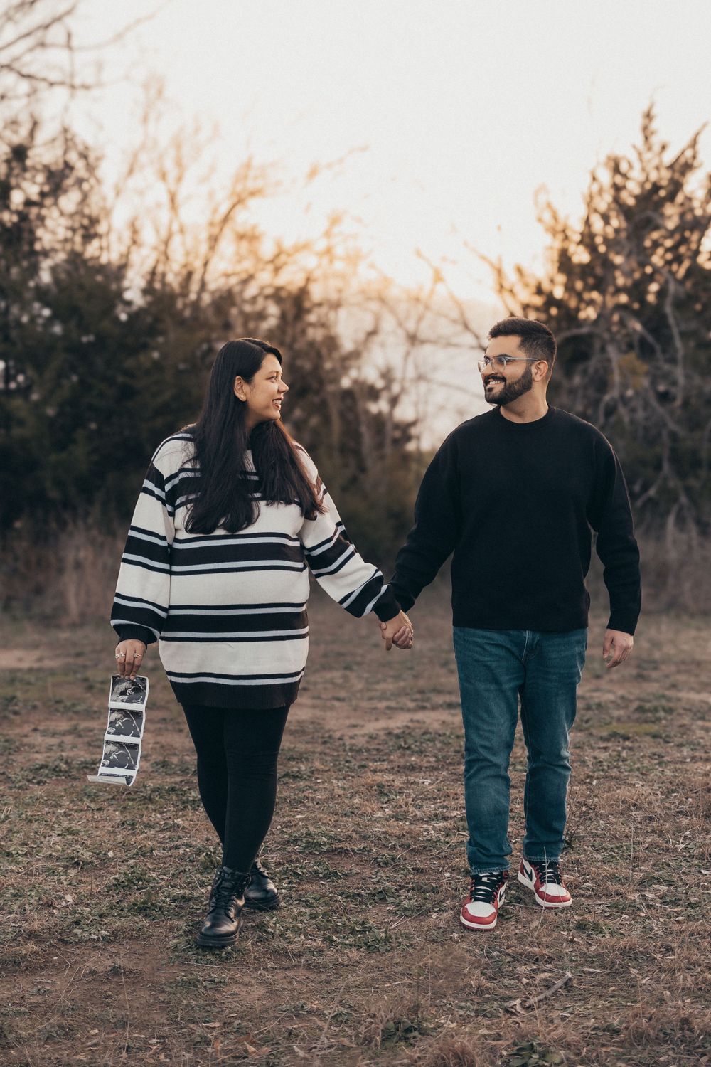A couple walks hand in hand through a field at sunset while holding ultrasound photos.