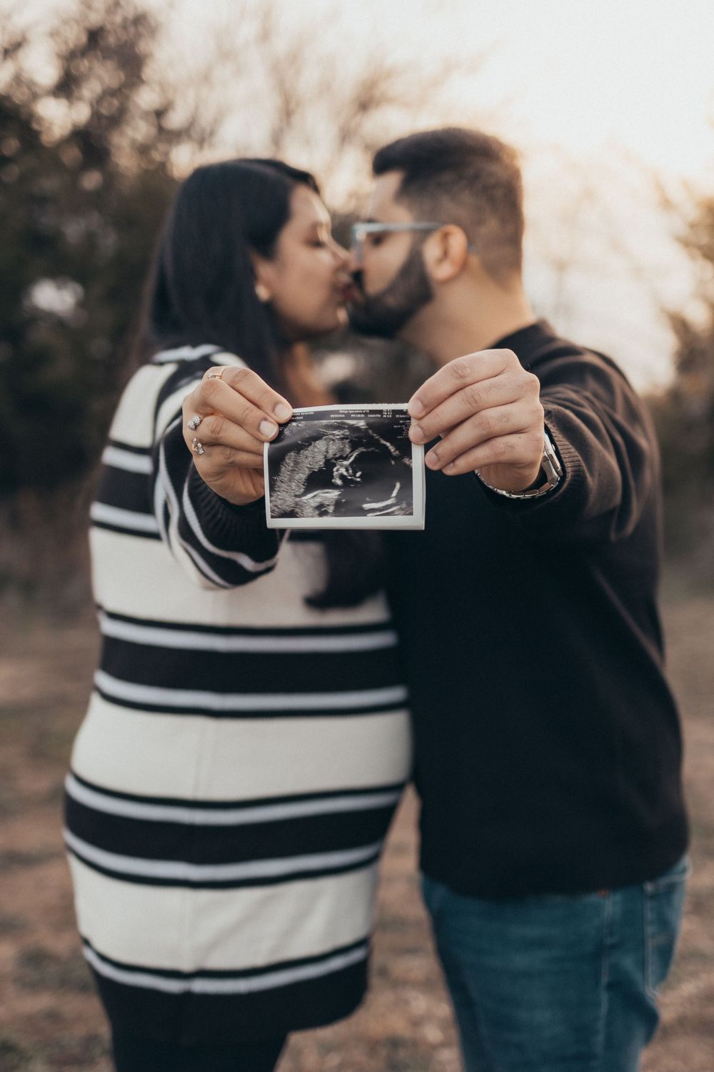 A couple kisses while holding up an ultrasound photo during sunset.