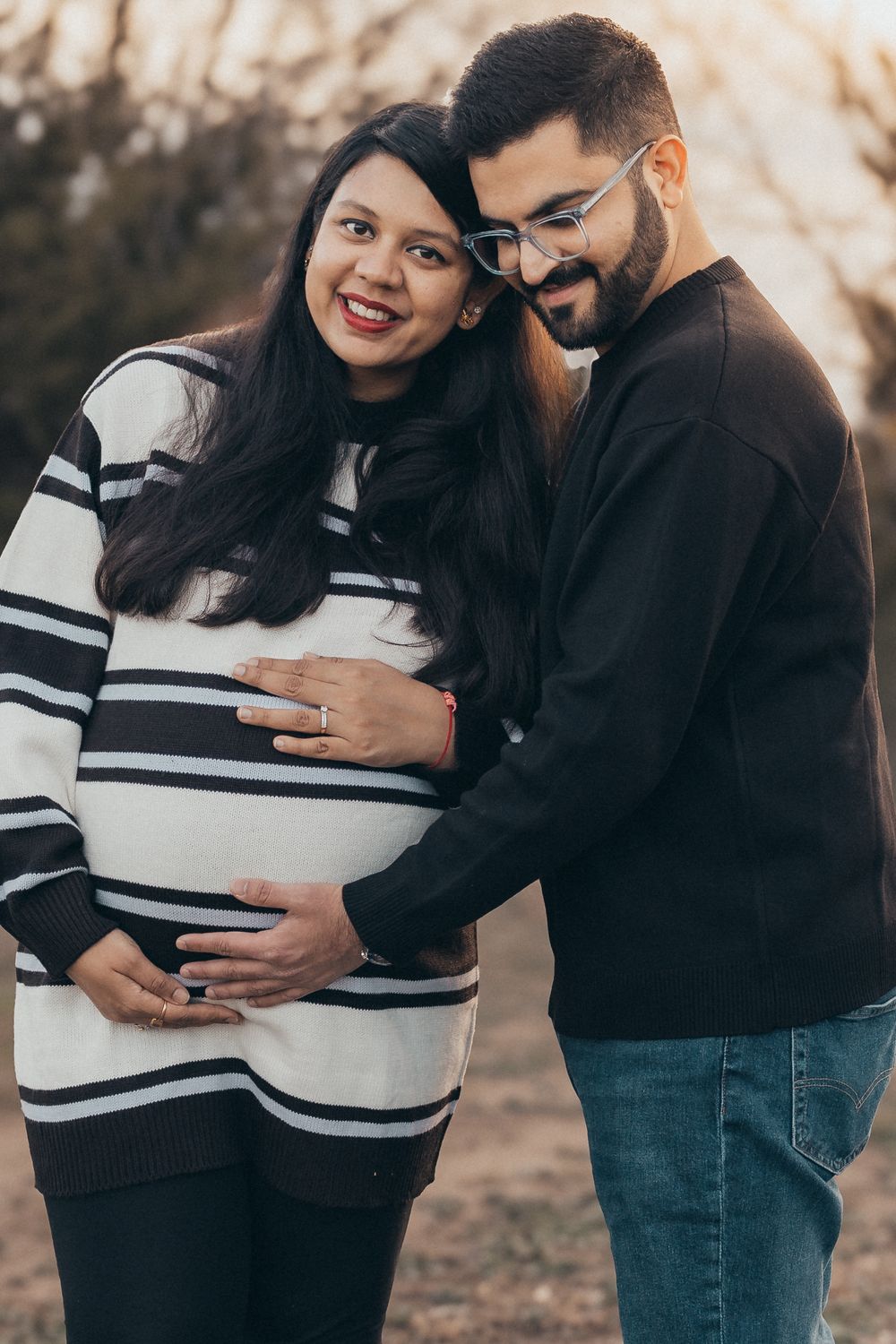 Expecting couple embraces outdoors with hands on pregnant belly in winter clothing at sunset.