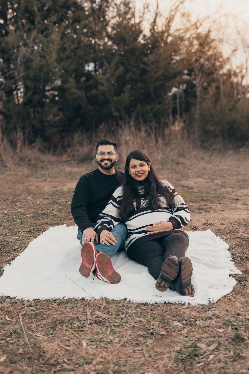 Couple sits on white blanket outdoors enjoying maternity photoshoot at dusk.