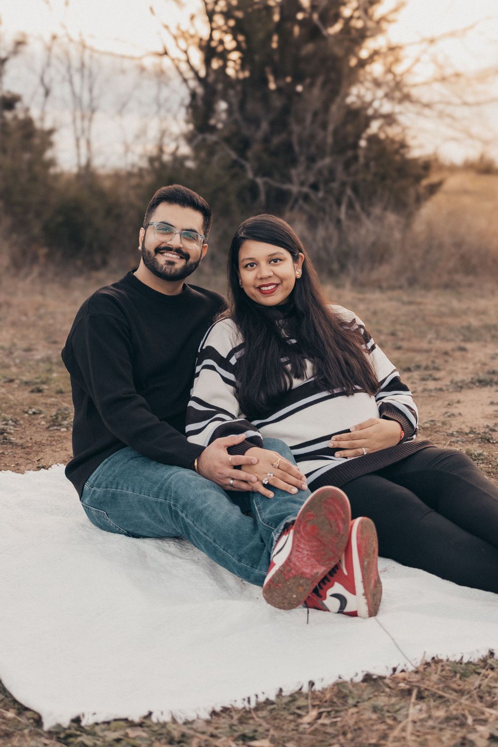Couple relaxes on white blanket during outdoor maternity session at golden hour.
