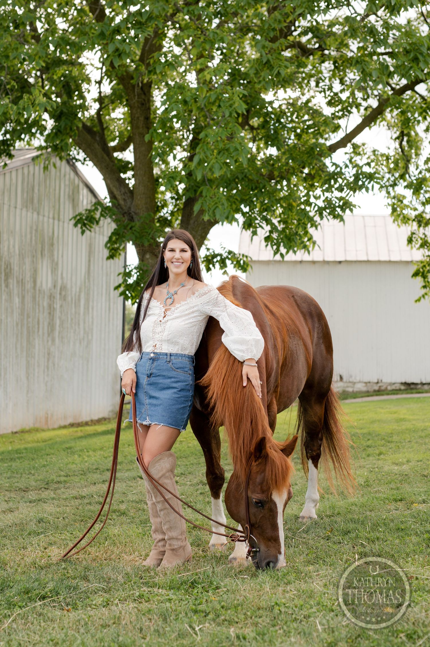 Woman in white top and denim skirt stands beside grazing brown horse near white barn wall.