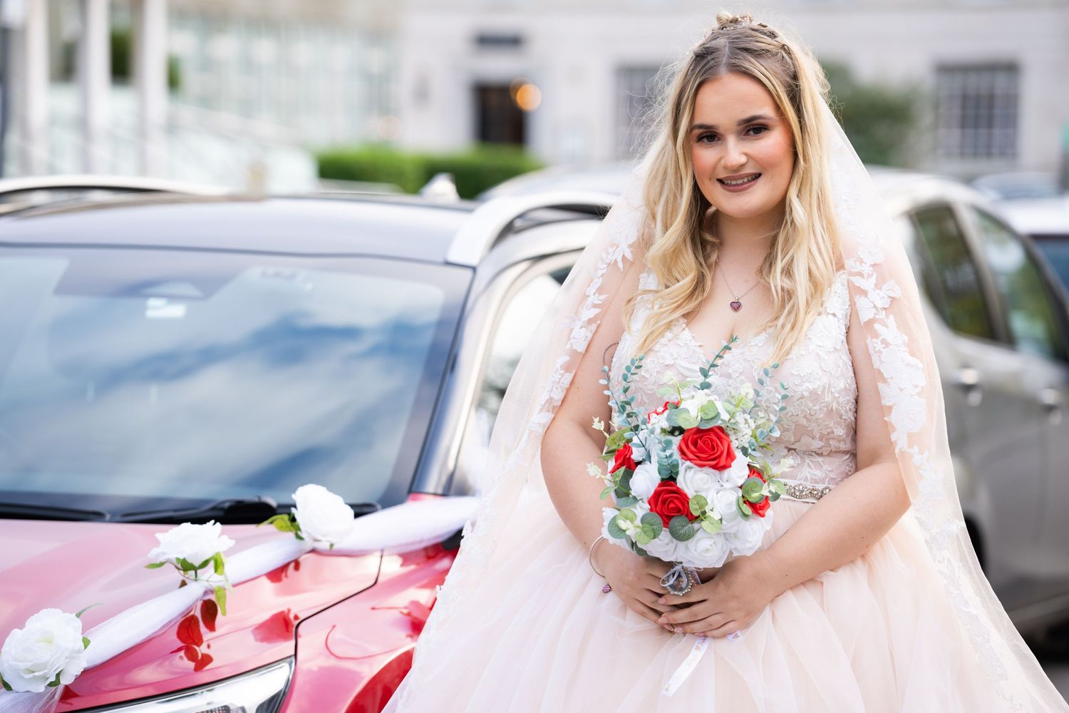 A bride in a flowing white dress holds a red and white bouquet next to a decorated wedding car.