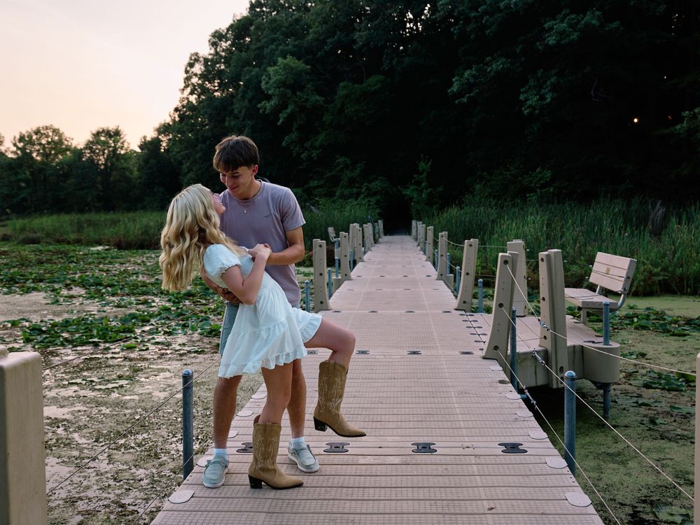 A couple shares a romantic moment on a wooden boardwalk surrounded by lily pads and nature at sunset.