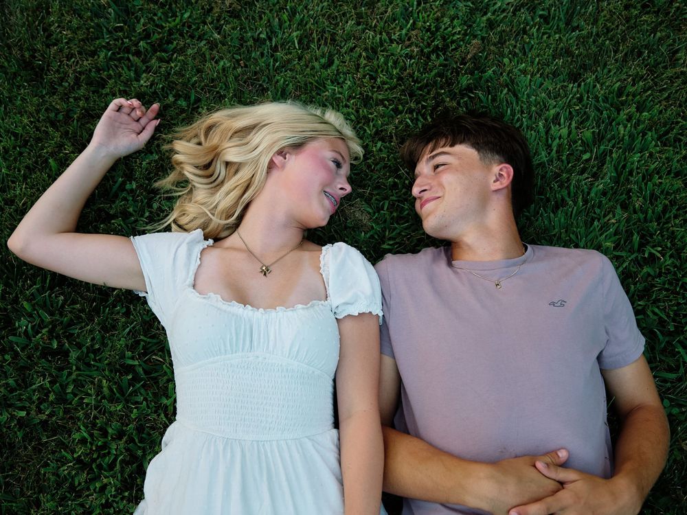 Couple lying together on green grass wearing white and gray shirts in a romantic pose.