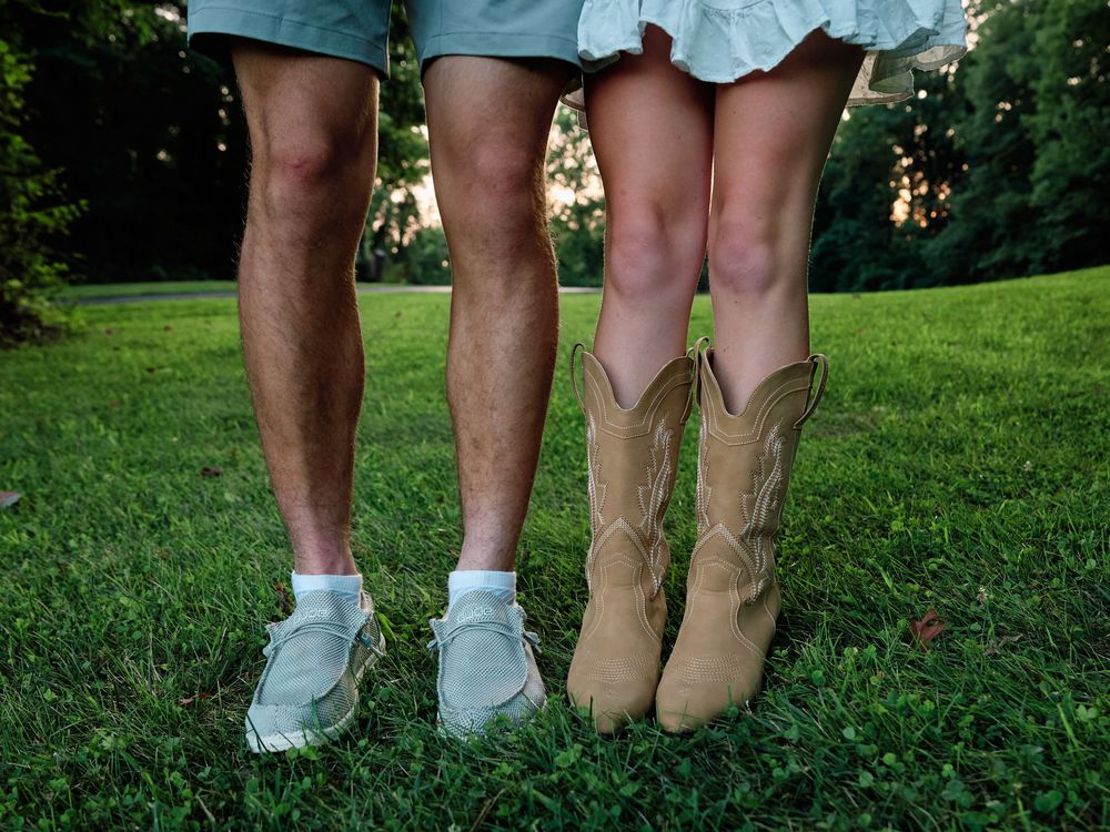 Two pairs of legs wearing different colored shoes stand together on green grass in a park setting.