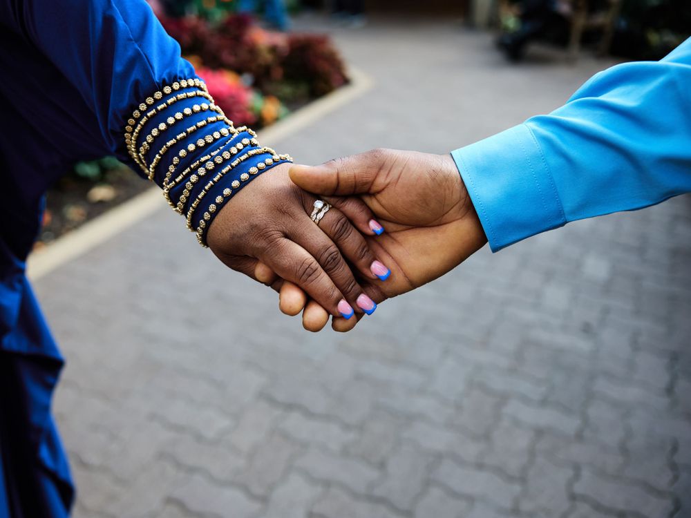 Two hands hold each other, with one wearing blue bangles and purple nail polish, on a blurred paved background.