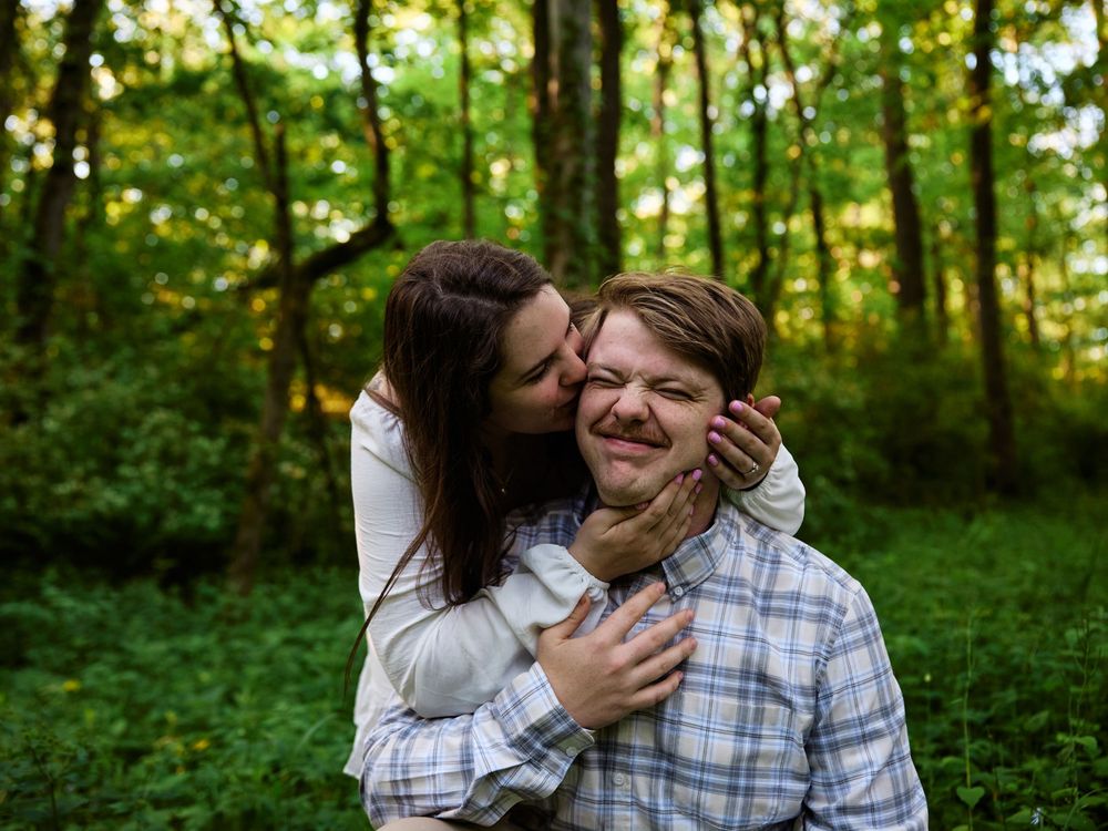 A playful couple embraces and laughs together in a sunlit forest setting.