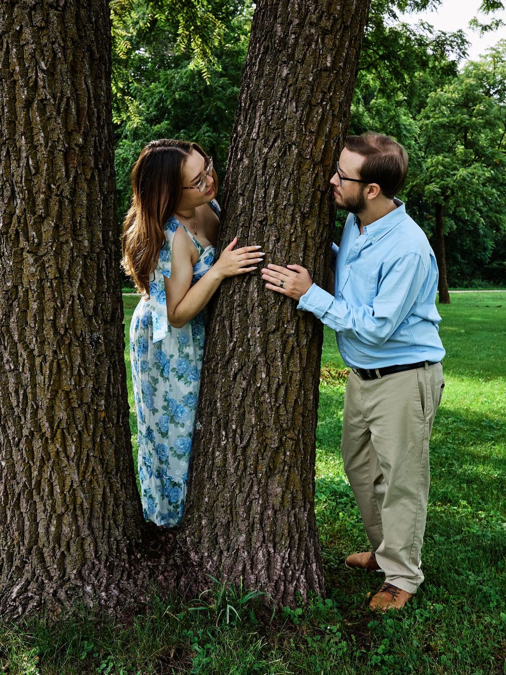 A couple playfully interacts on opposite sides of a large tree trunk in a green summer park setting.