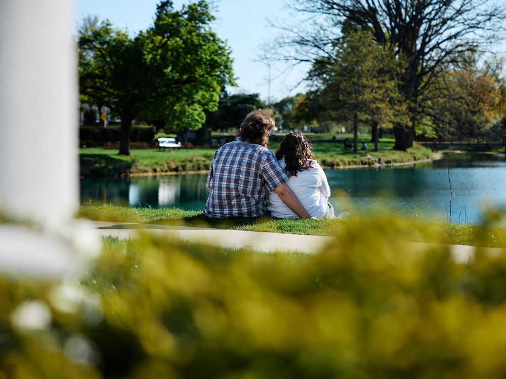 A couple sits on the shore of a tranquil lake surrounded by trees and greenery on a sunny day.