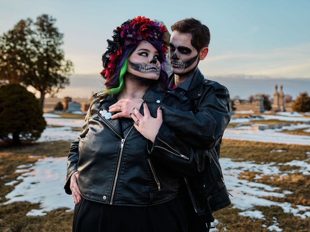 Couple in Day of the Dead skull makeup and leather jackets pose together in snowy winter landscape at sunset.