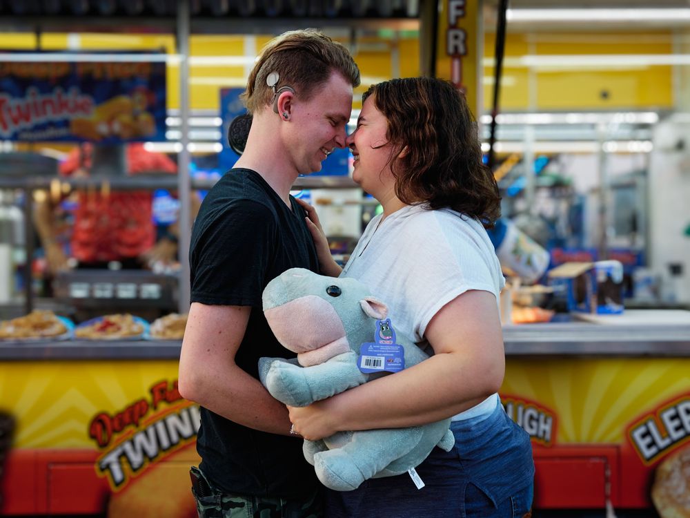 A couple shares a tender moment at a carnival or fair with colorful concession stands in the background.