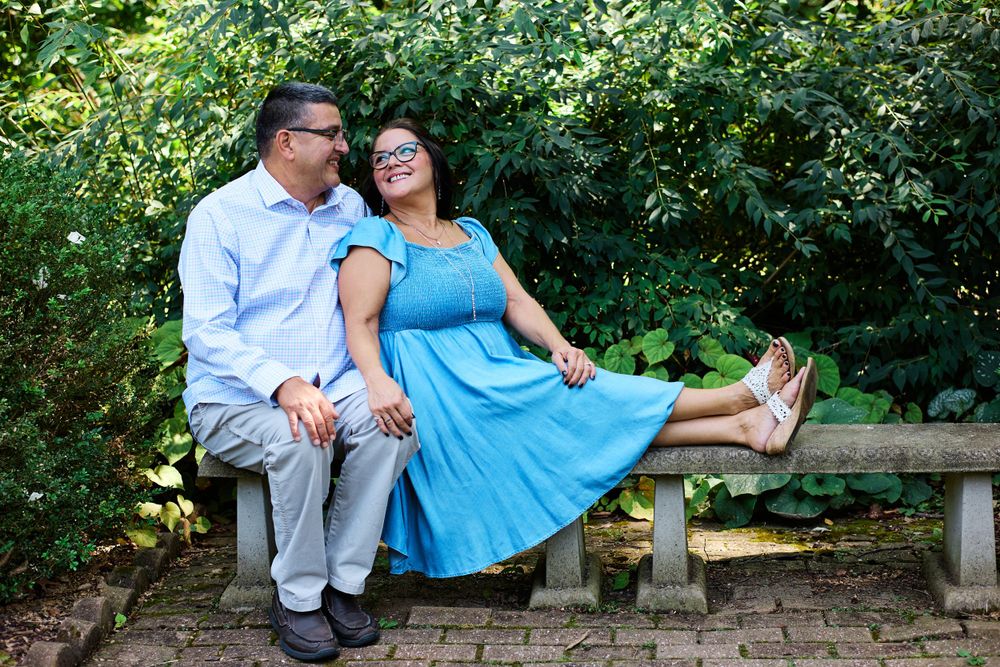 A couple shares a joyful moment while sitting on a stone bench in a lush garden setting.