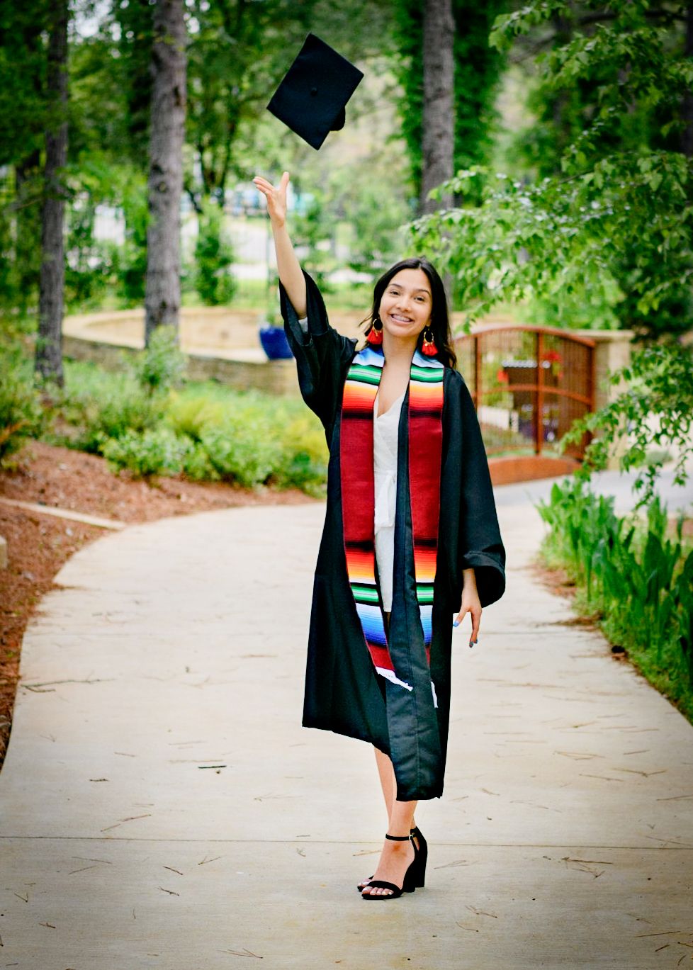 Graduate in black cap and gown celebrating by tossing cap in wooded outdoor setting.