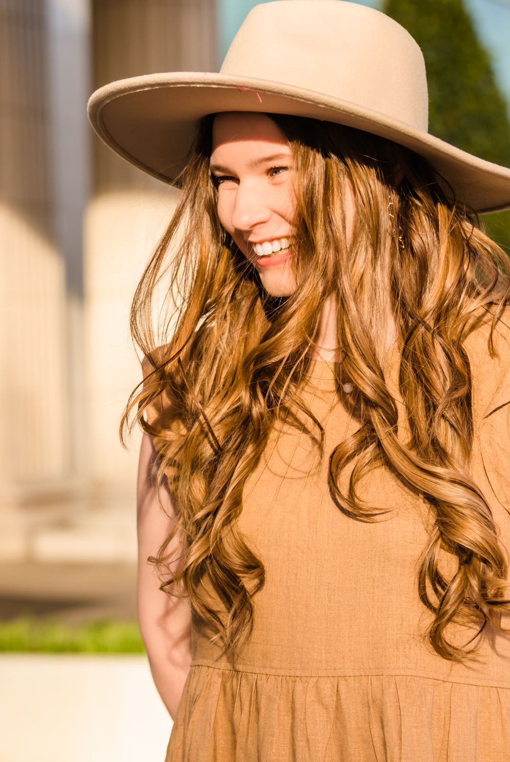 Person in tan dress and wide-brimmed hat smiles in outdoor portrait.