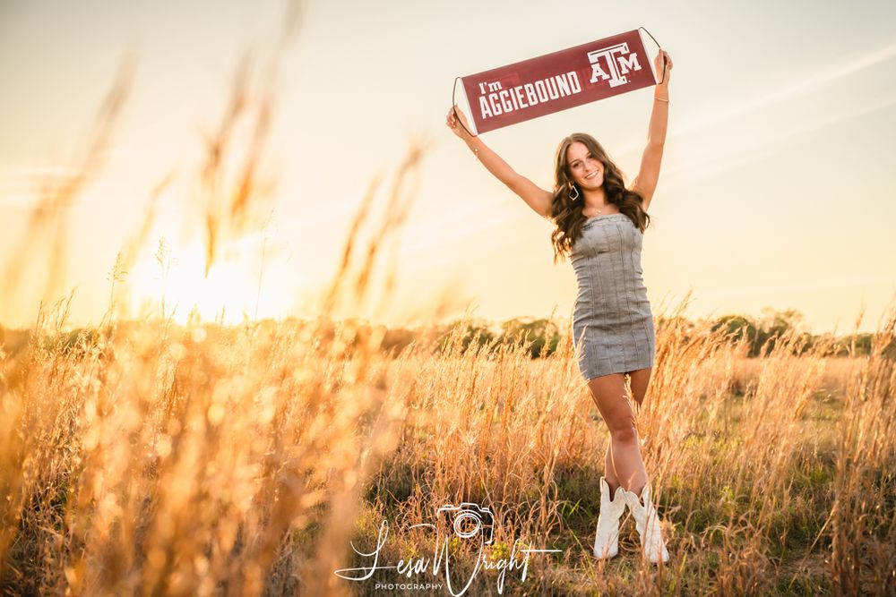 A figure holds a Texas A&M sign while standing in a golden wheat field at sunset wearing boots and a gray dress.