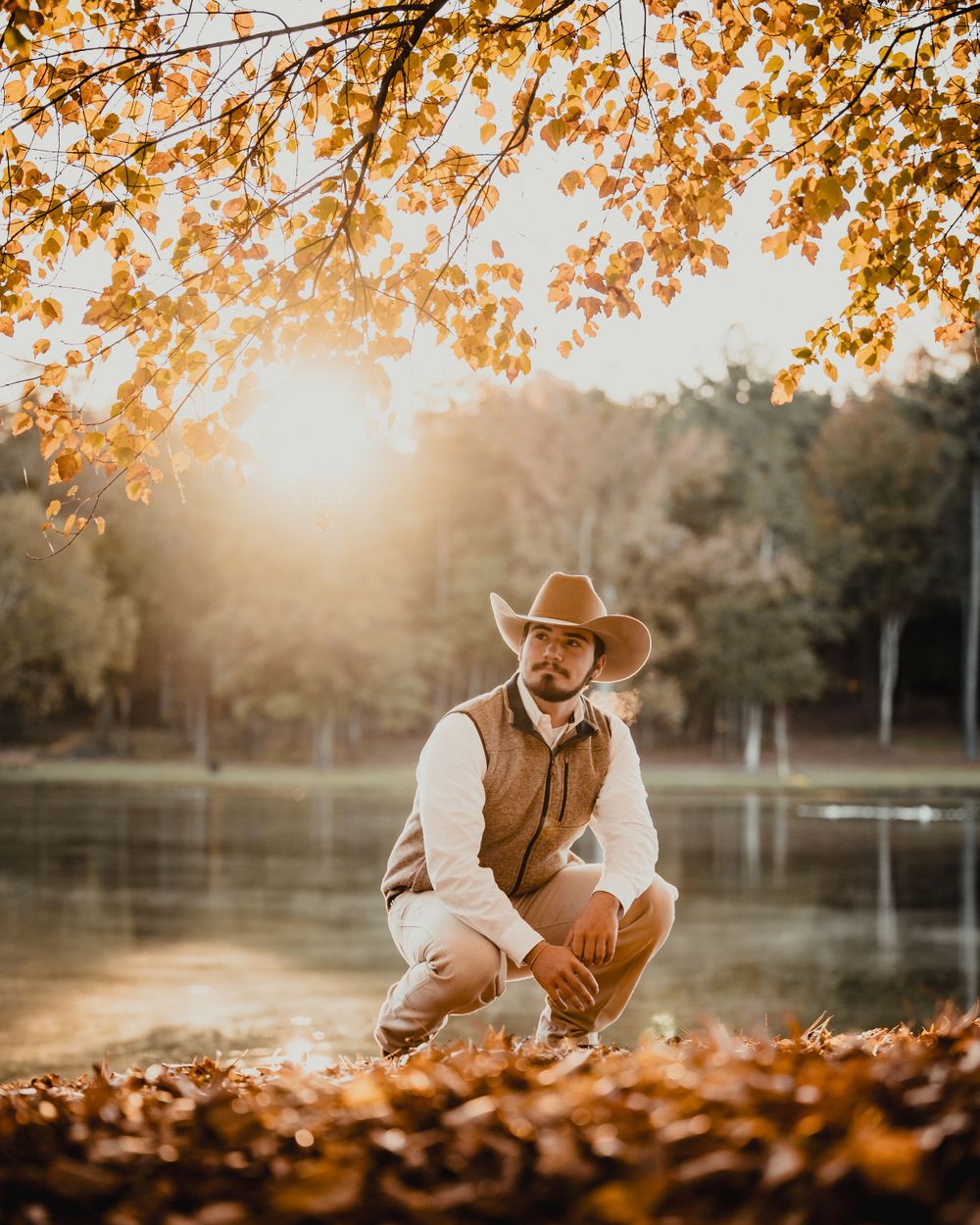 A figure in cowboy attire crouches by a lake at sunset with golden autumn leaves overhead.