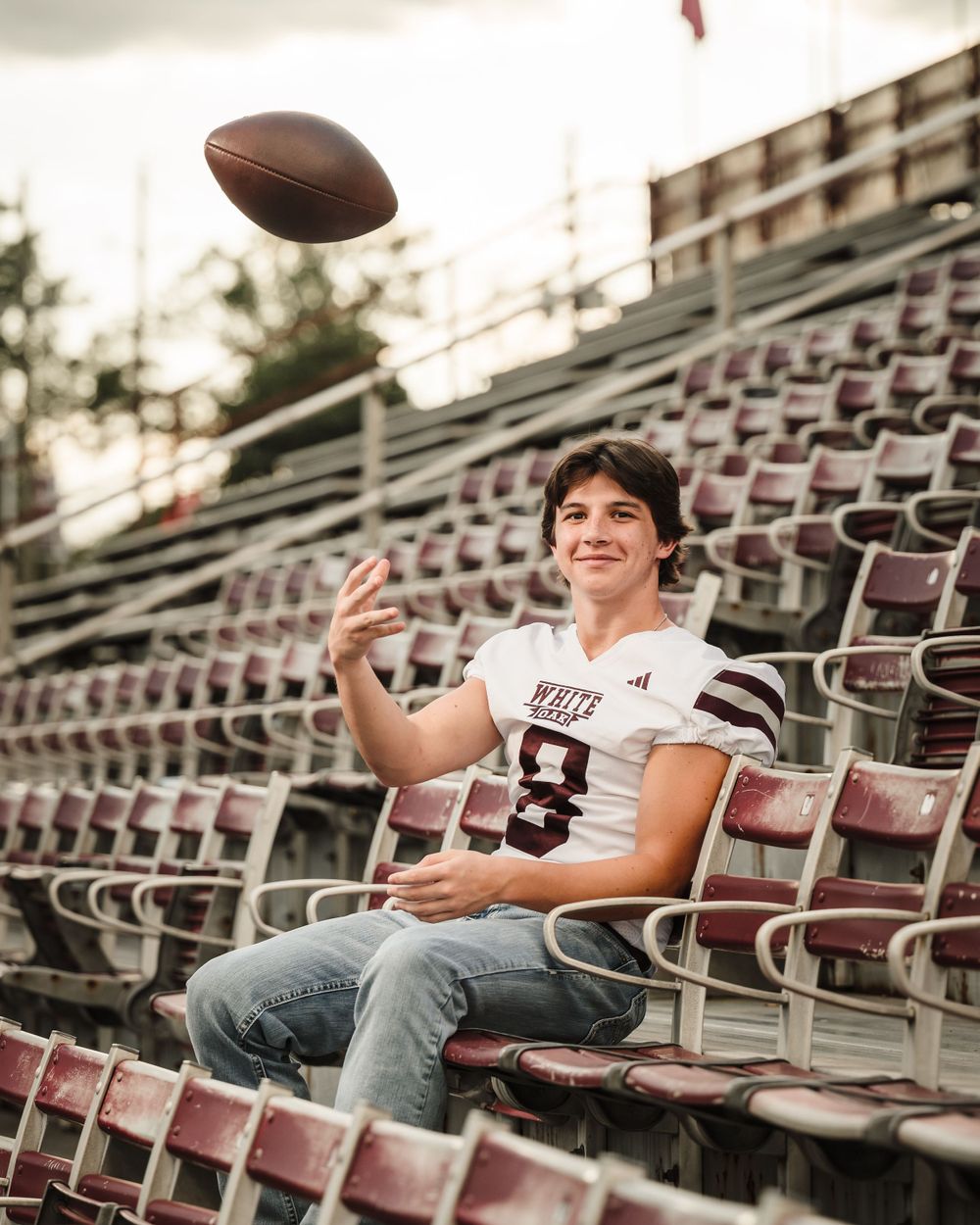 Football player tossing ball while sitting in empty stadium bleachers at sunset.