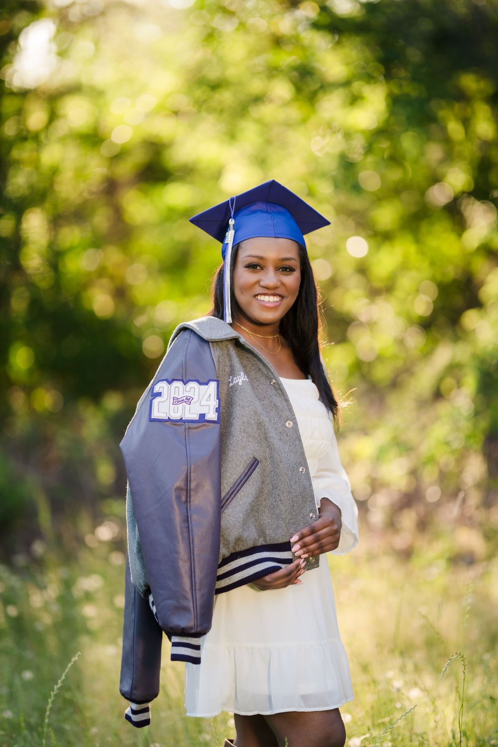 Graduate posing outdoors in a varsity jacket with white dress and blue cap and gown.