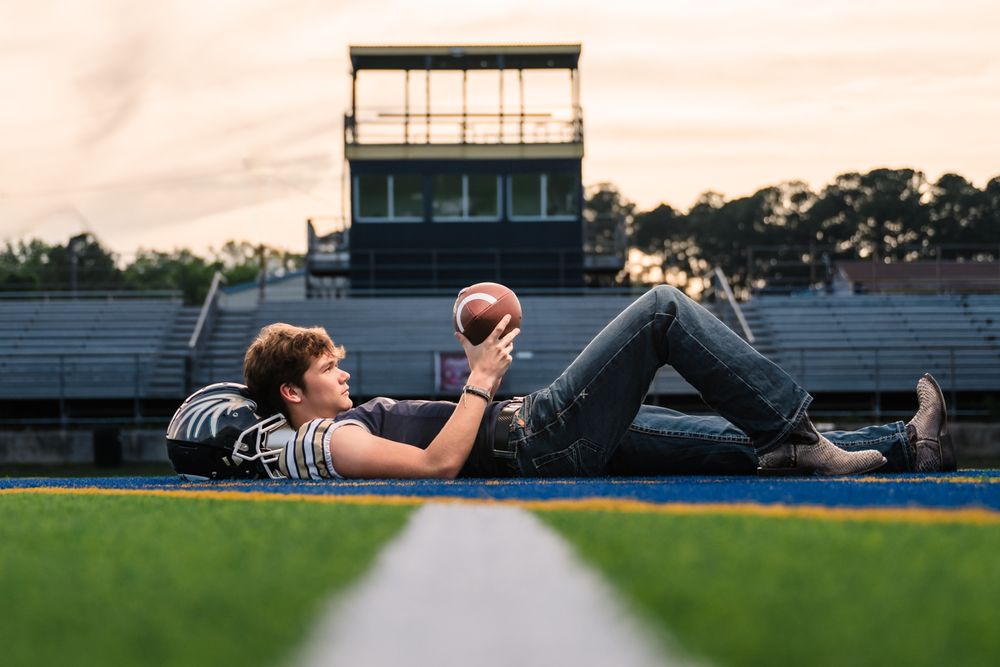 Teen boy lying on football field holding a football at sunset, wearing jeans and shoulder pads.