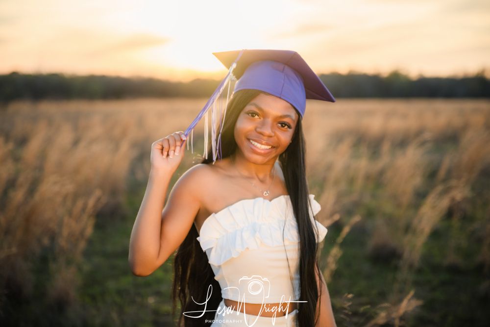 Graduate wearing purple cap and white dress stands in golden field at sunset.