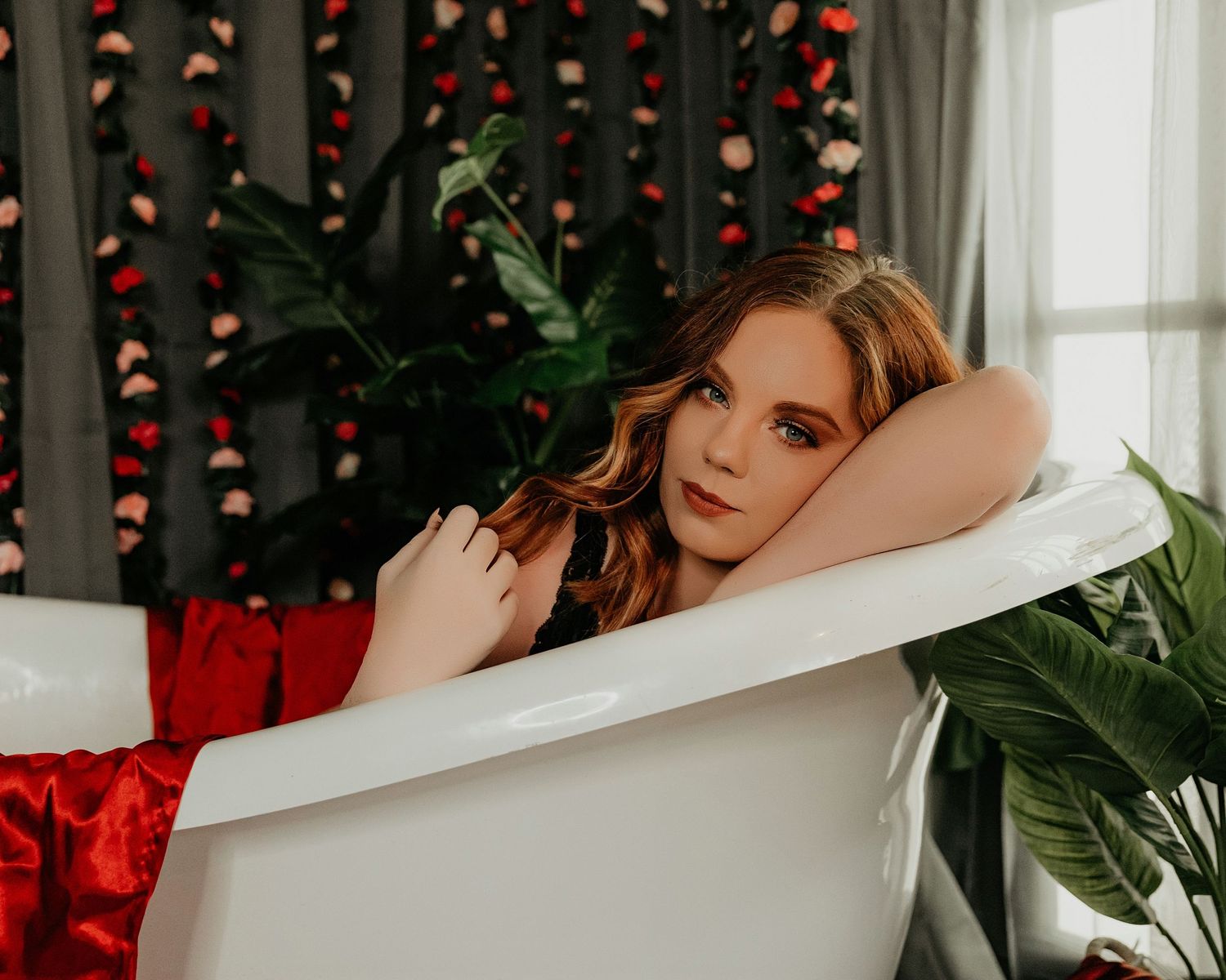 Boudoir portrait in white bathtub with red decor and tropical plants.