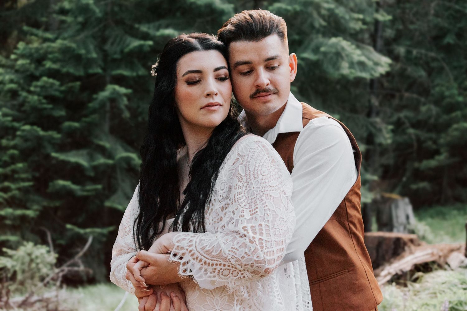 A striking couple posed in a deep green forest, showcasing timeless love with modern rustic outfits and romantic editorial posing.