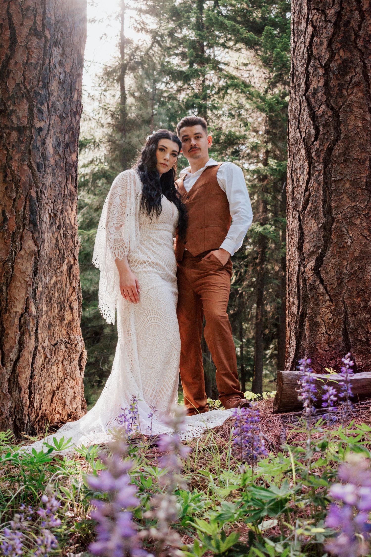 A dreamy engagement photo of a couple embracing under tall pine trees surrounded by vibrant purple wildflowers in a lush forest near Seattle