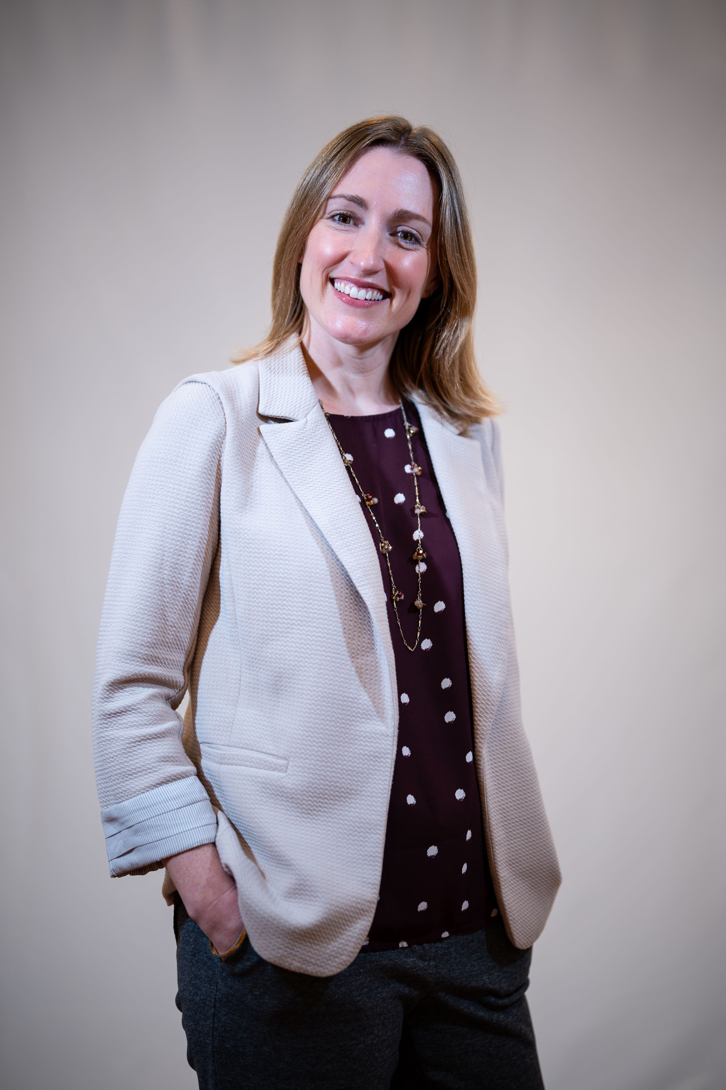 Professional portrait of someone wearing a cream blazer and burgundy top with a delicate necklace against neutral background.