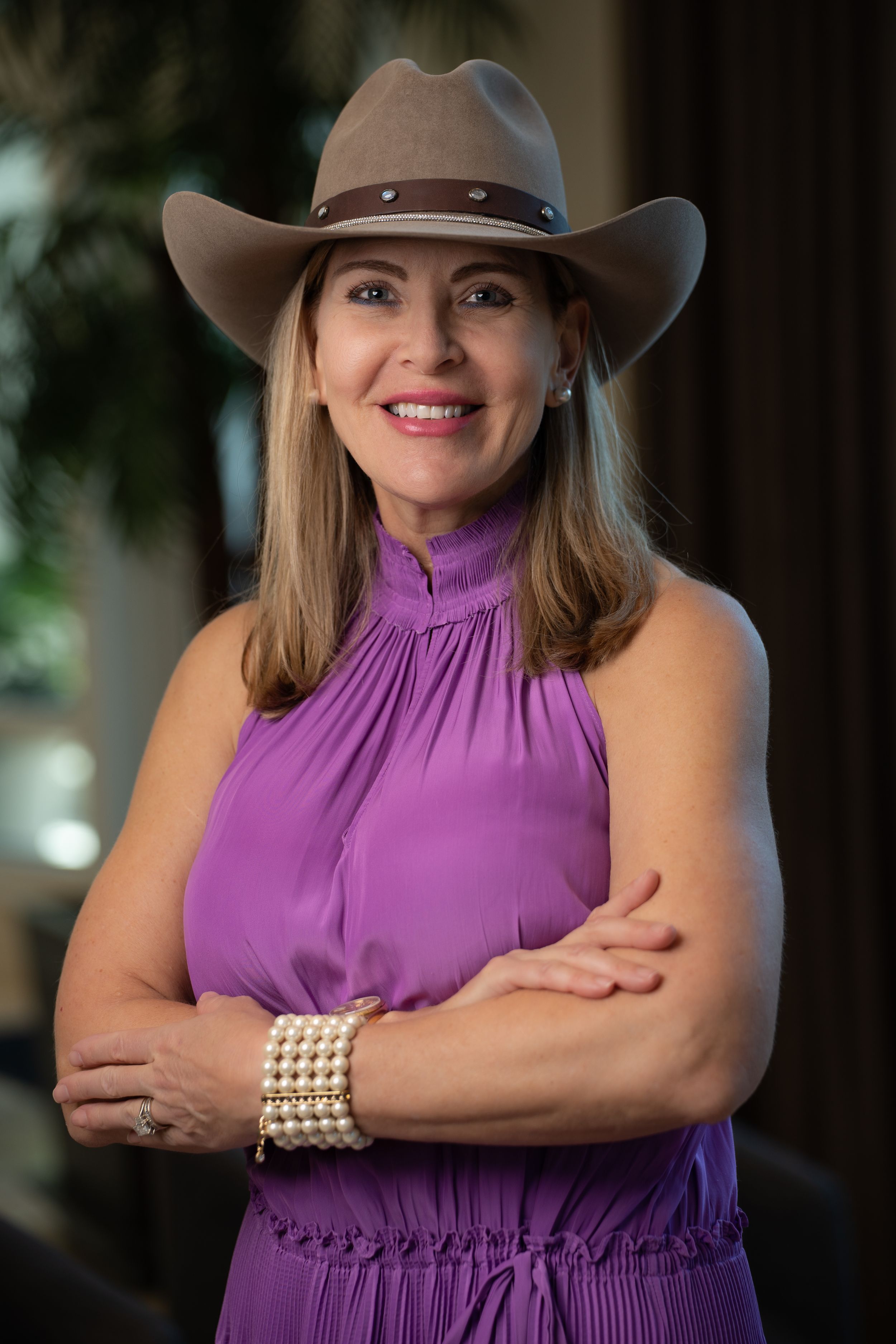 A person wearing a brown cowboy hat and purple sleeveless dress poses with arms crossed and pearl bracelet.