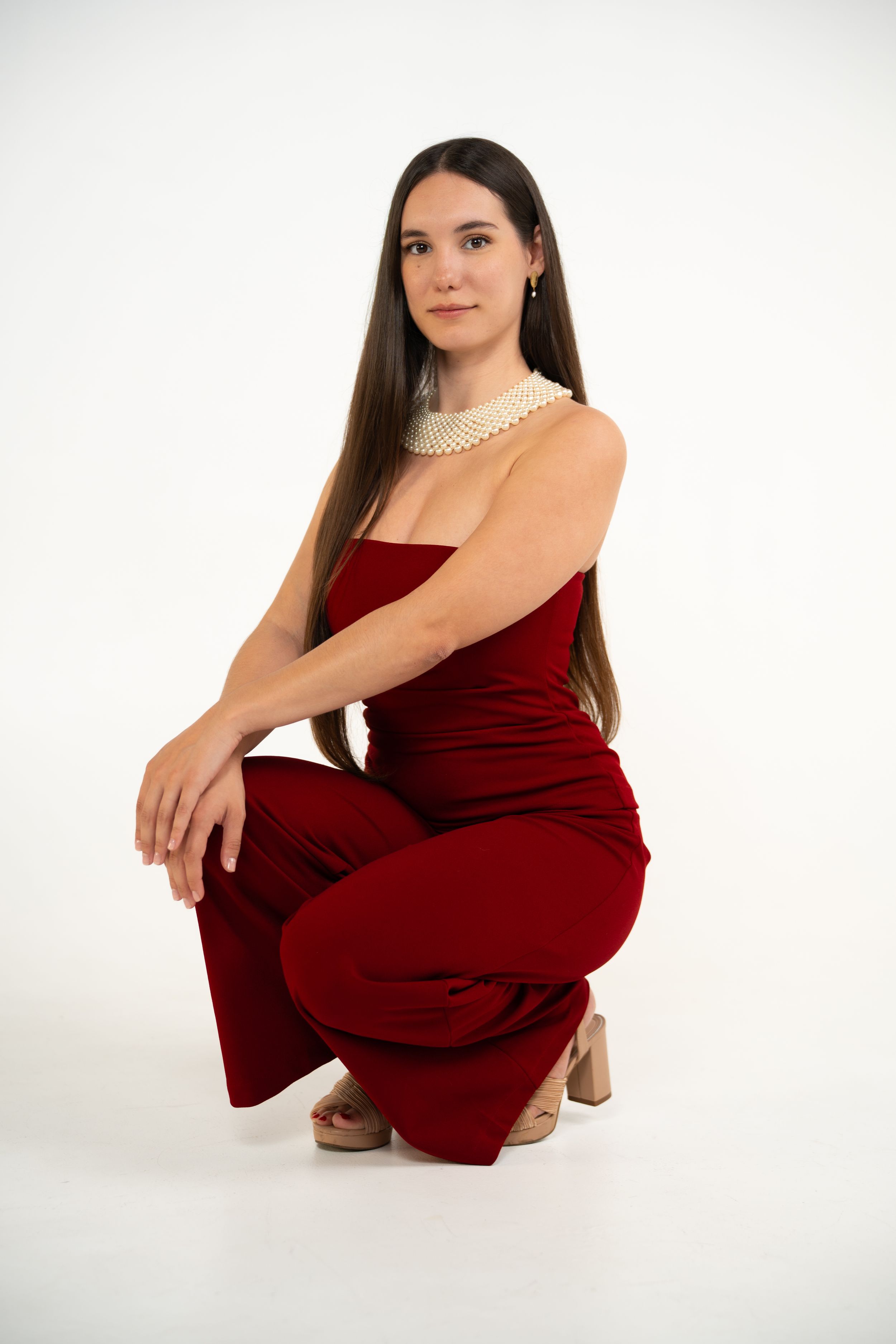 A person in a red strapless jumpsuit and pearl necklace crouches elegantly against a white studio backdrop.