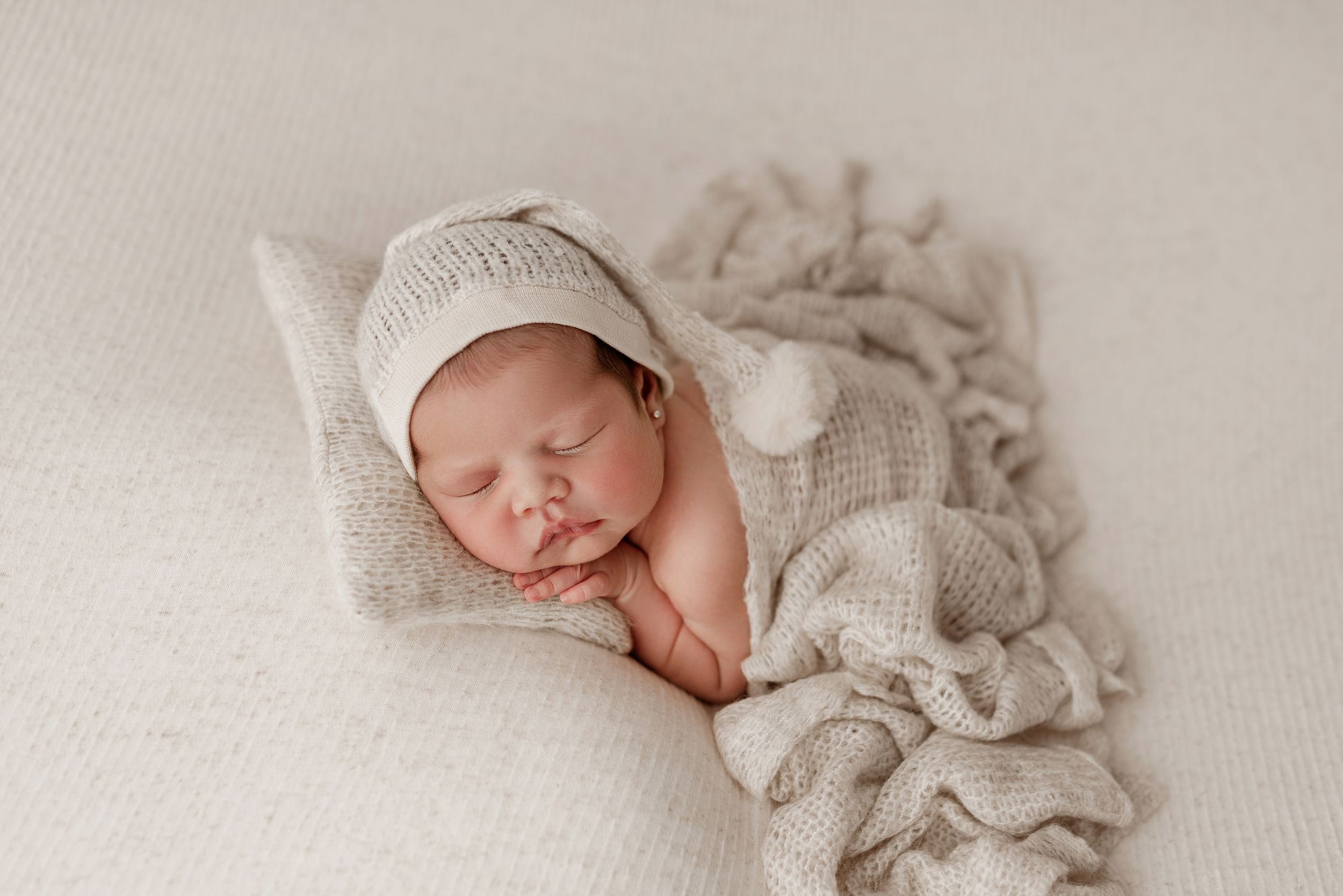 A peaceful sleeping newborn baby wrapped in soft beige fabrics and wearing a white knit bonnet on textured backdrop.