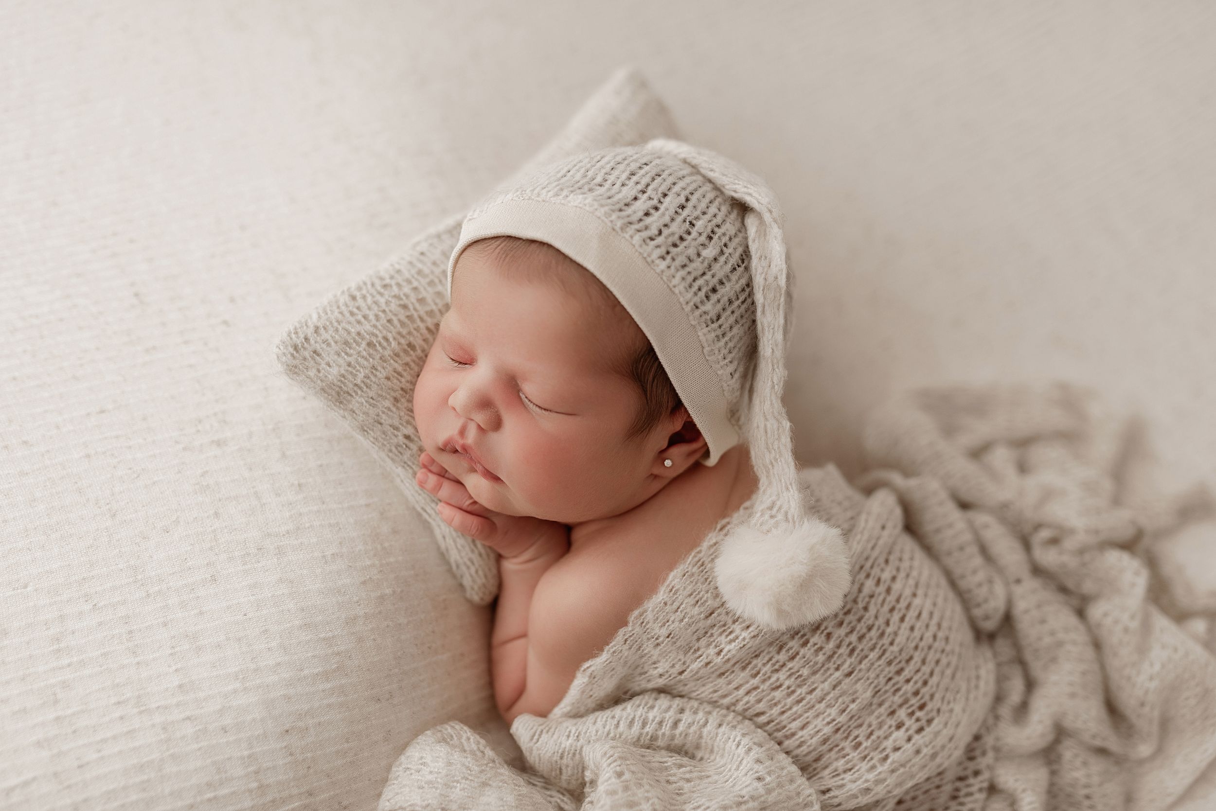 Sleeping newborn wearing soft knitted beanie and wrapped in cozy white textured blanket during studio photo session.