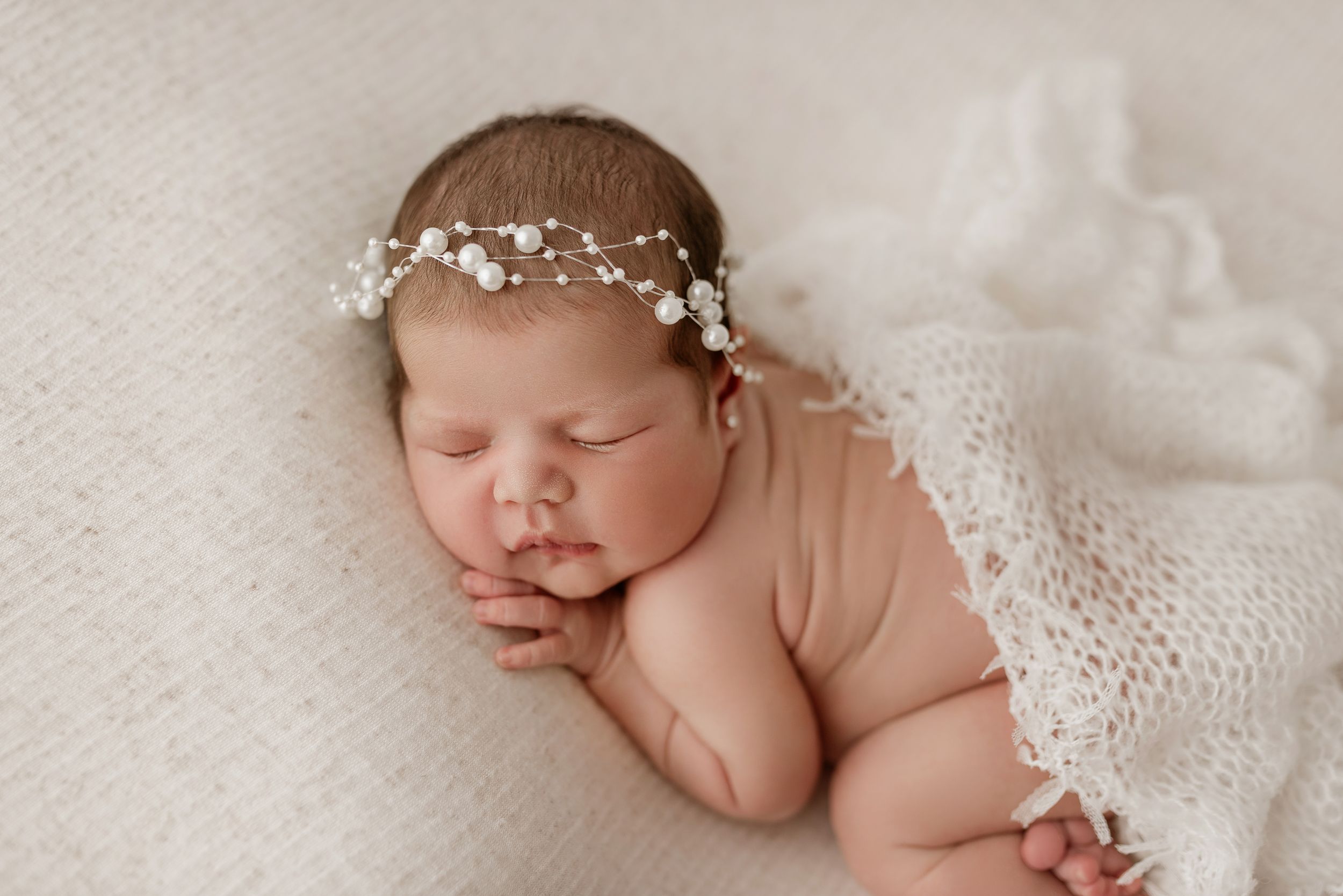 Sleeping newborn wearing delicate white floral crown rests peacefully on soft white textured blanket.
