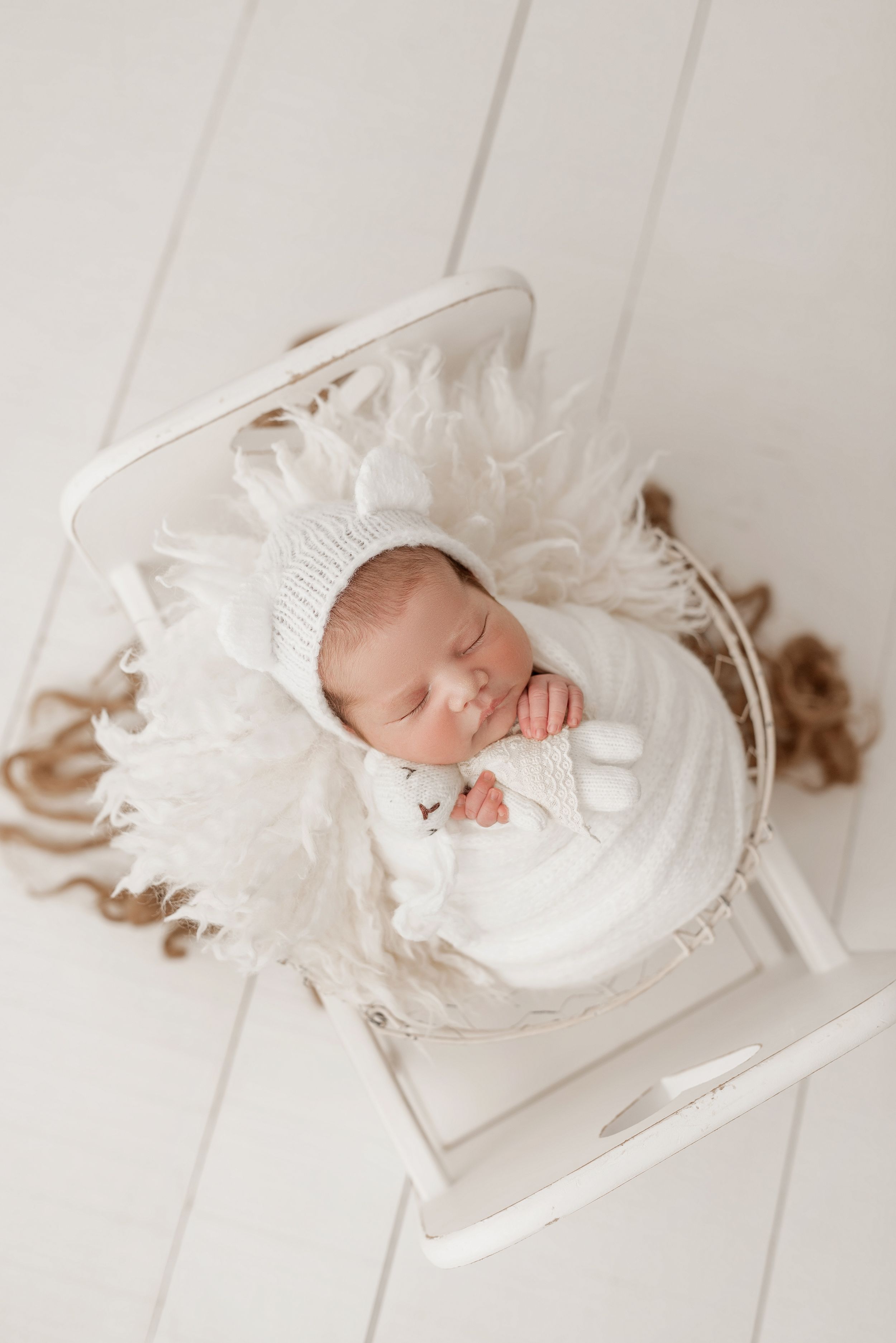 A sleeping newborn in white swaddle and bonnet rests peacefully on a white wooden surface with soft floral accents.