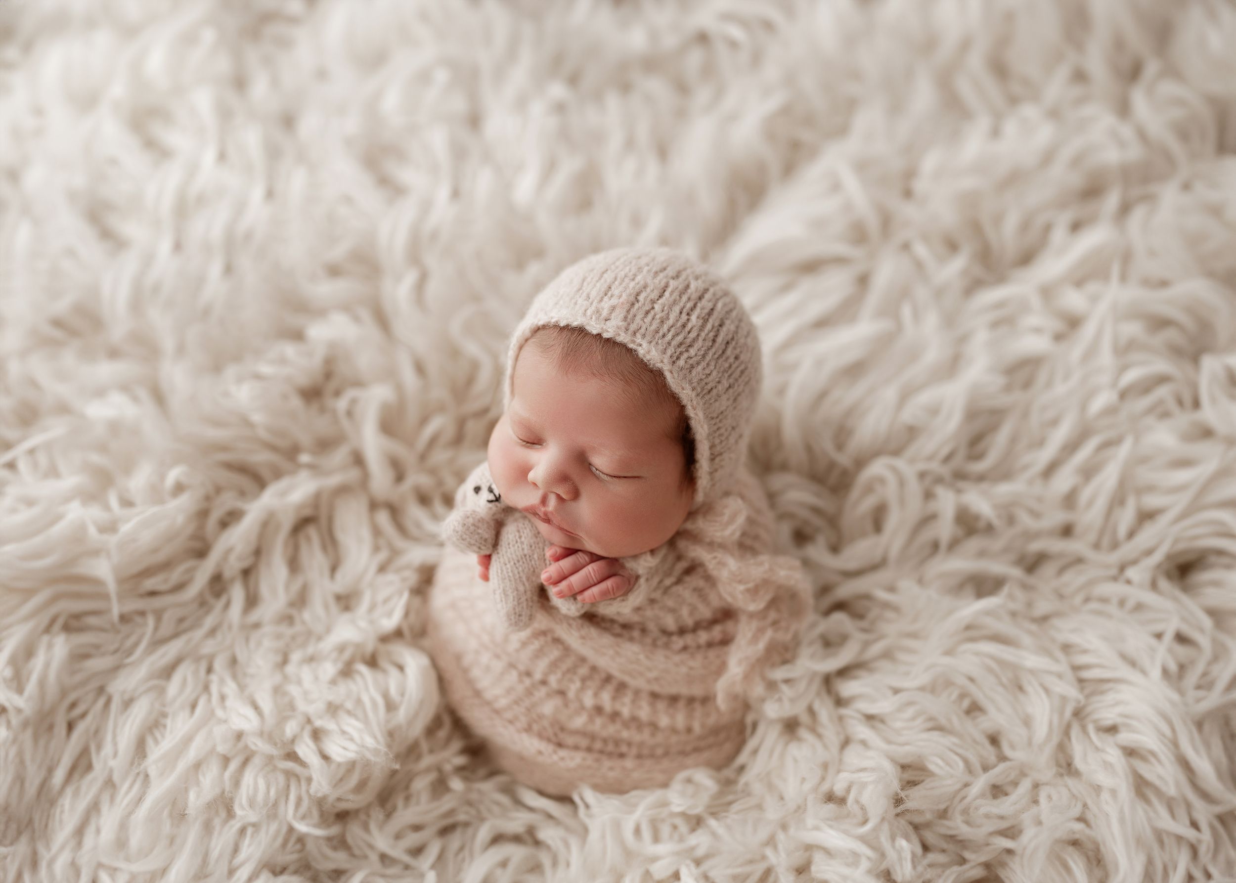 A newborn sleeps on cream-colored fluffy fabric while wearing a knitted beige bonnet and sweater.