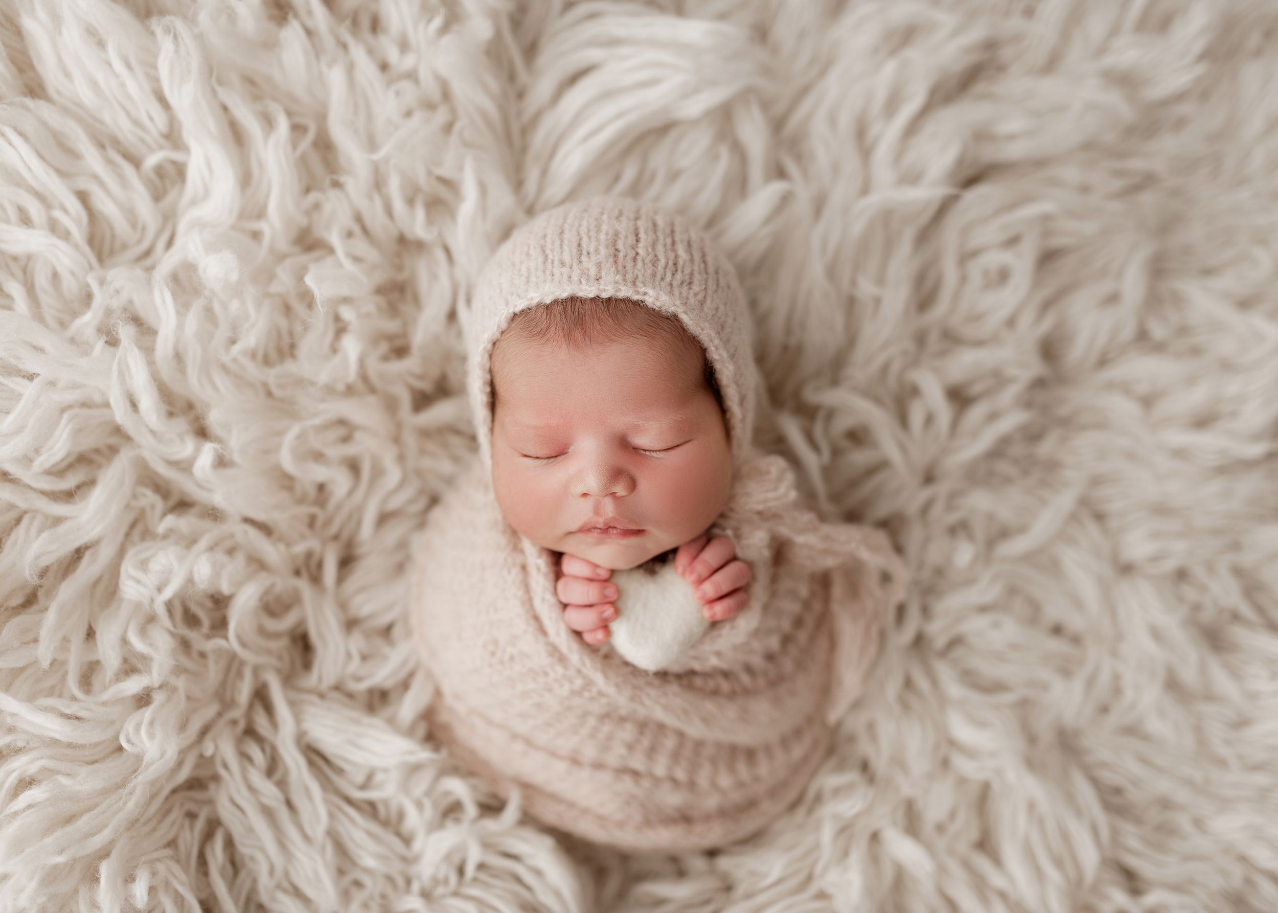 Sleeping newborn baby wrapped in soft beige knit blanket resting on fluffy white fur backdrop.