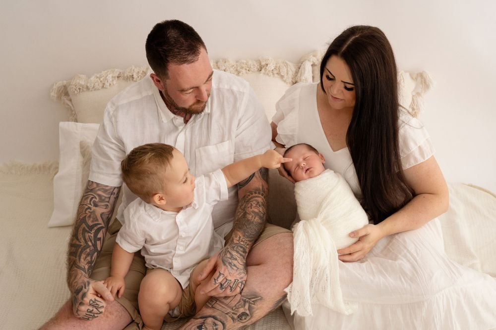 A family in white sits together on a bed, with parents lovingly gazing at their children in a tender moment.