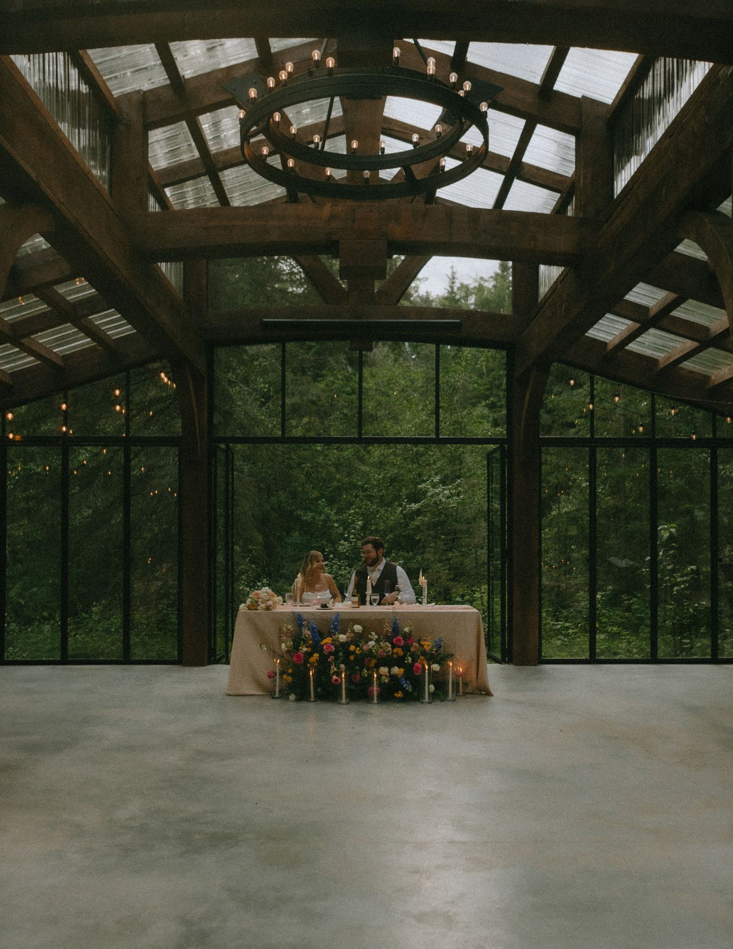 An elegant reception table setup under a glass ceiling conservatory with chandelier and forest views.