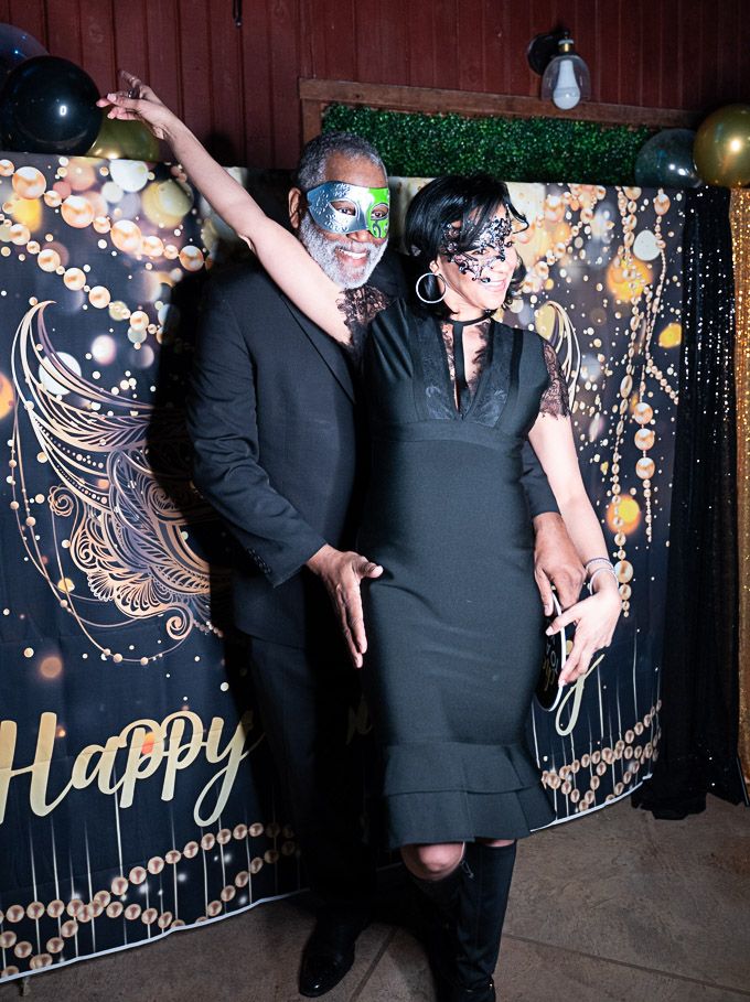 Pixs By Tina captures a couple in formal black attire posing against a decorative wall with gold accents at a celebration event