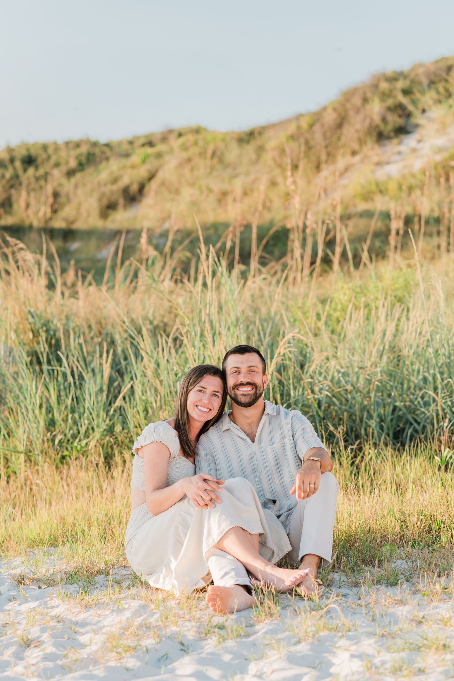 A couple sits together on a sandy beach with tall grass and hills in the background during golden hour.