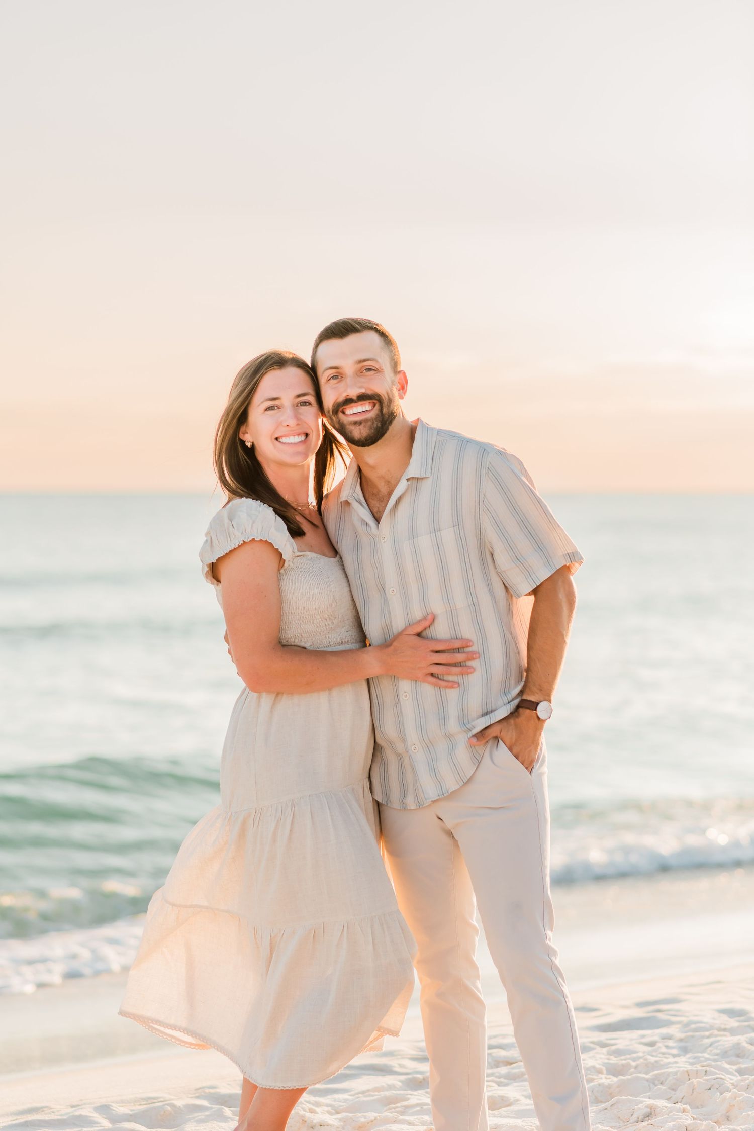 A couple shares a joyful moment on the beach at sunset, wearing light-colored casual outfits against the ocean view.