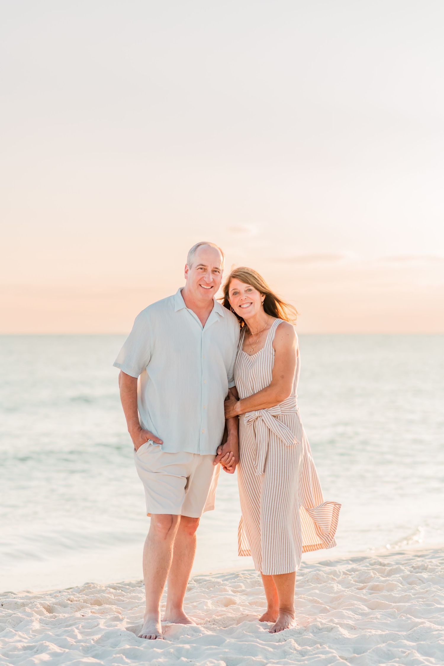 Couple in light colored outfits sharing romantic moment on white sandy beach at sunset.