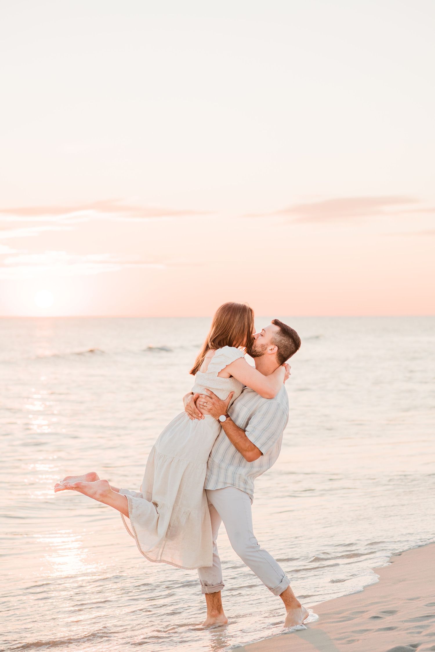 A couple embraces and dances together on a beach at sunset with soft pink sky overhead.