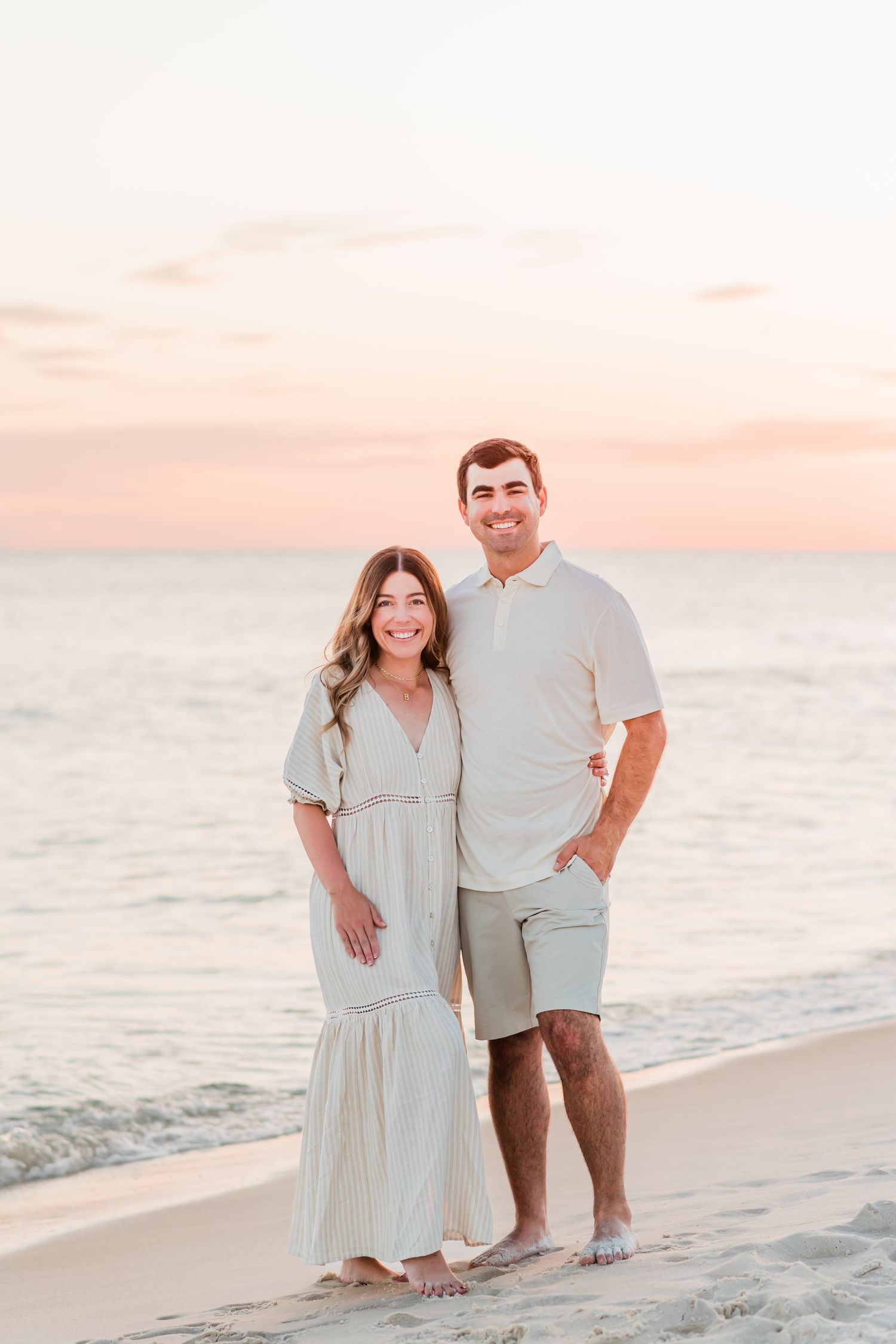 A couple stands together on a peaceful beach at sunset wearing coordinated light-colored outfits.