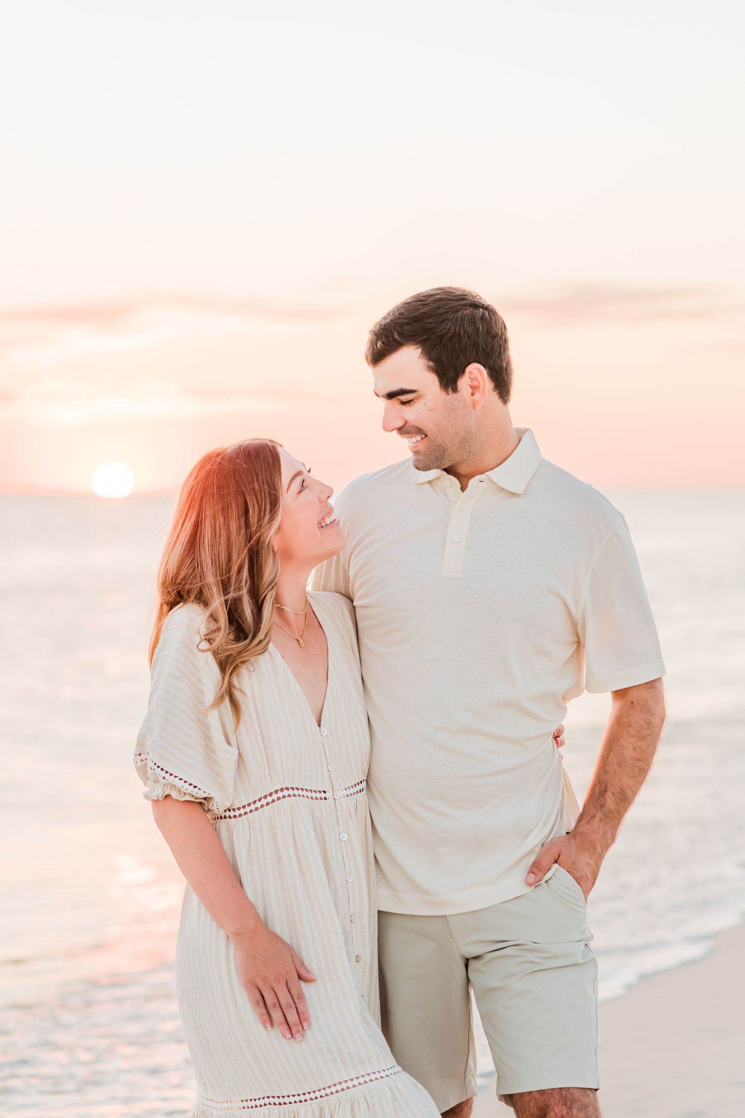 A romantic couple shares an intimate moment on the beach at sunset while wearing light colored summer clothing.